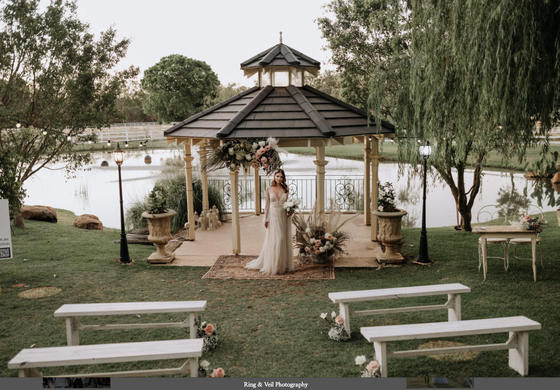 A bride in a wedding dress standing under a gazebo by a lake, with floral decorations and a grassy area with benches in the foreground.