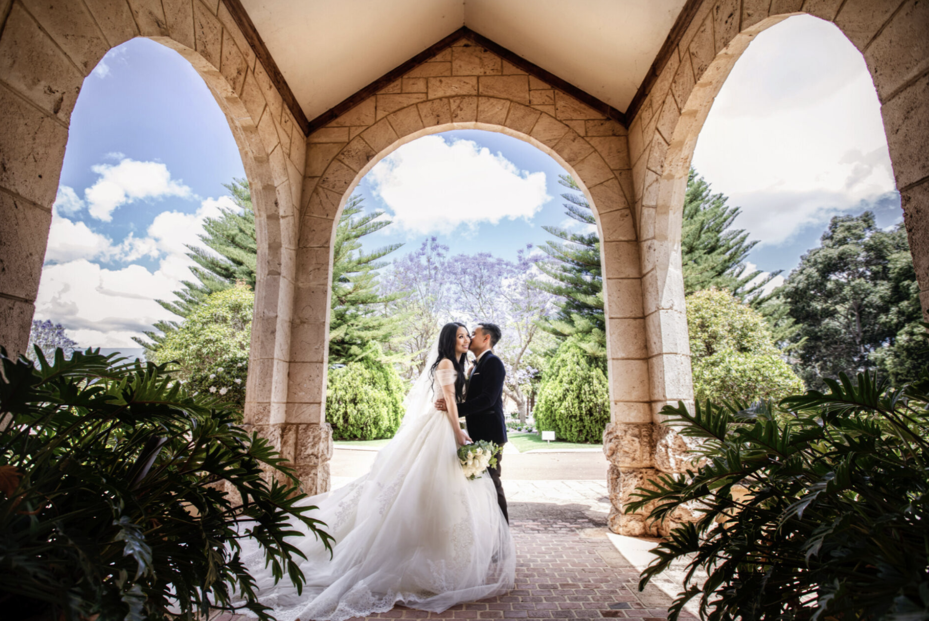 A bride and groom sharing a kiss under stone arches in a lush outdoor garden with trees and cloudy sky in the background.