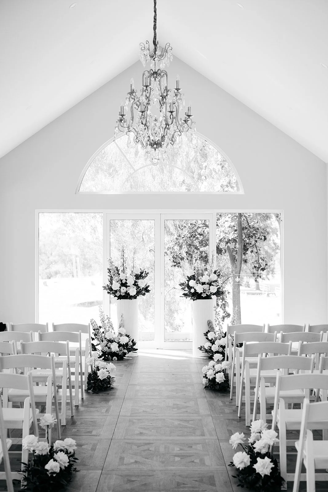 An elegant, bright indoor wedding chapel with a high vaulted ceiling, a chandelier, large windows, and floral arrangements along the aisle.