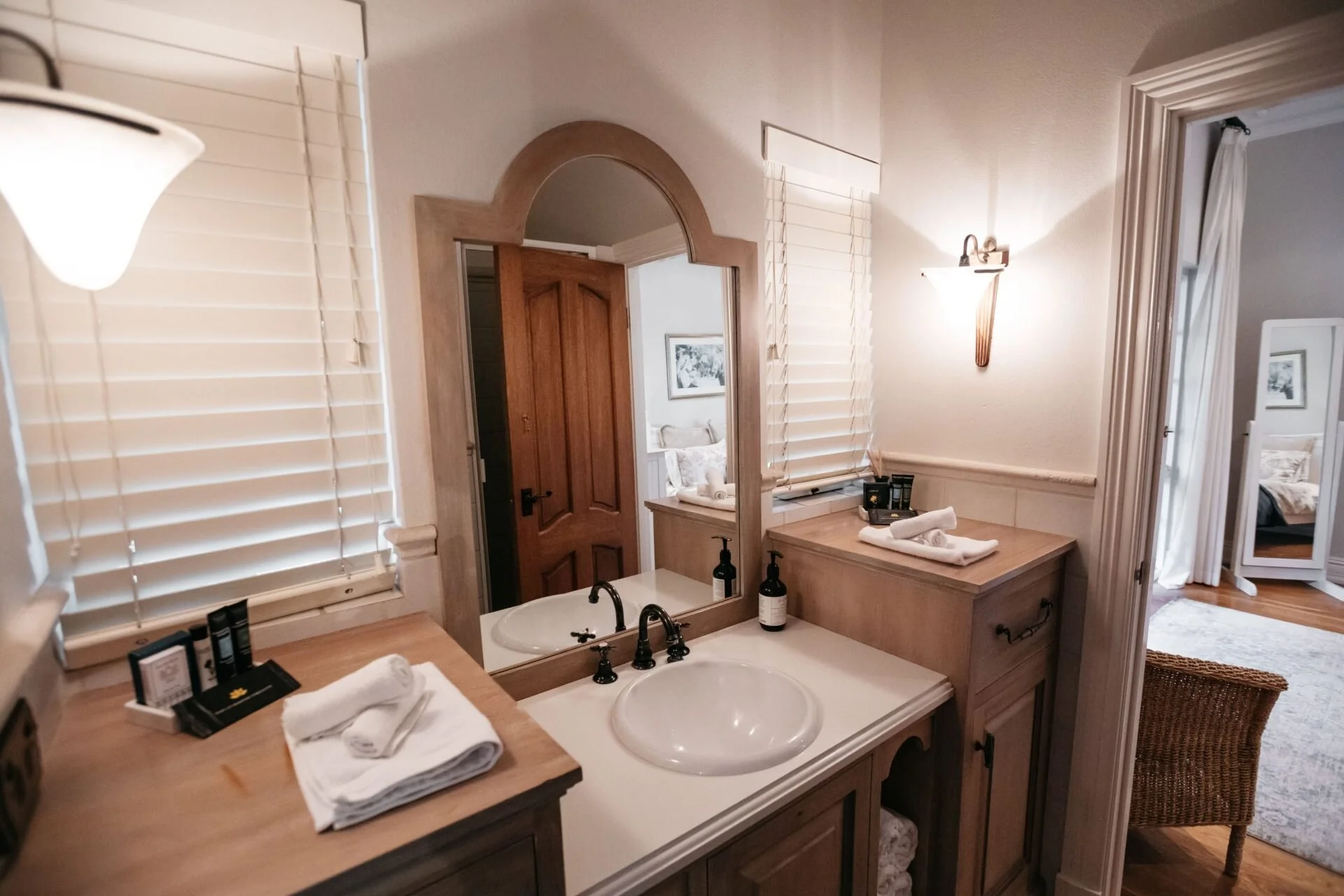 A bathroom with a wooden vanity, double sinks, a large mirror, white blinds on windows, wall-mounted lights, neatly folded towels, toiletries, and a view into a bedroom.