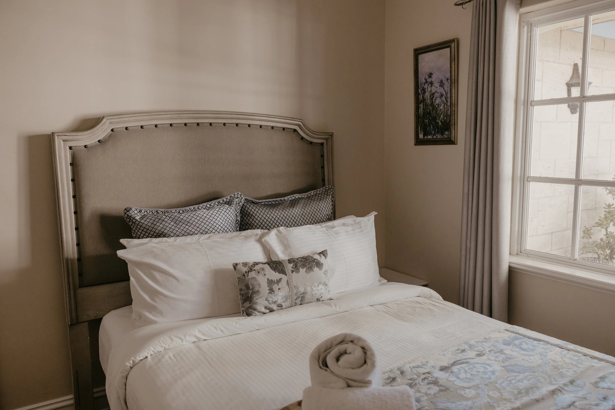 A neatly made bed with white sheets, decorative pillows, and a floral blanket, positioned beside a window with beige curtains and wall art.