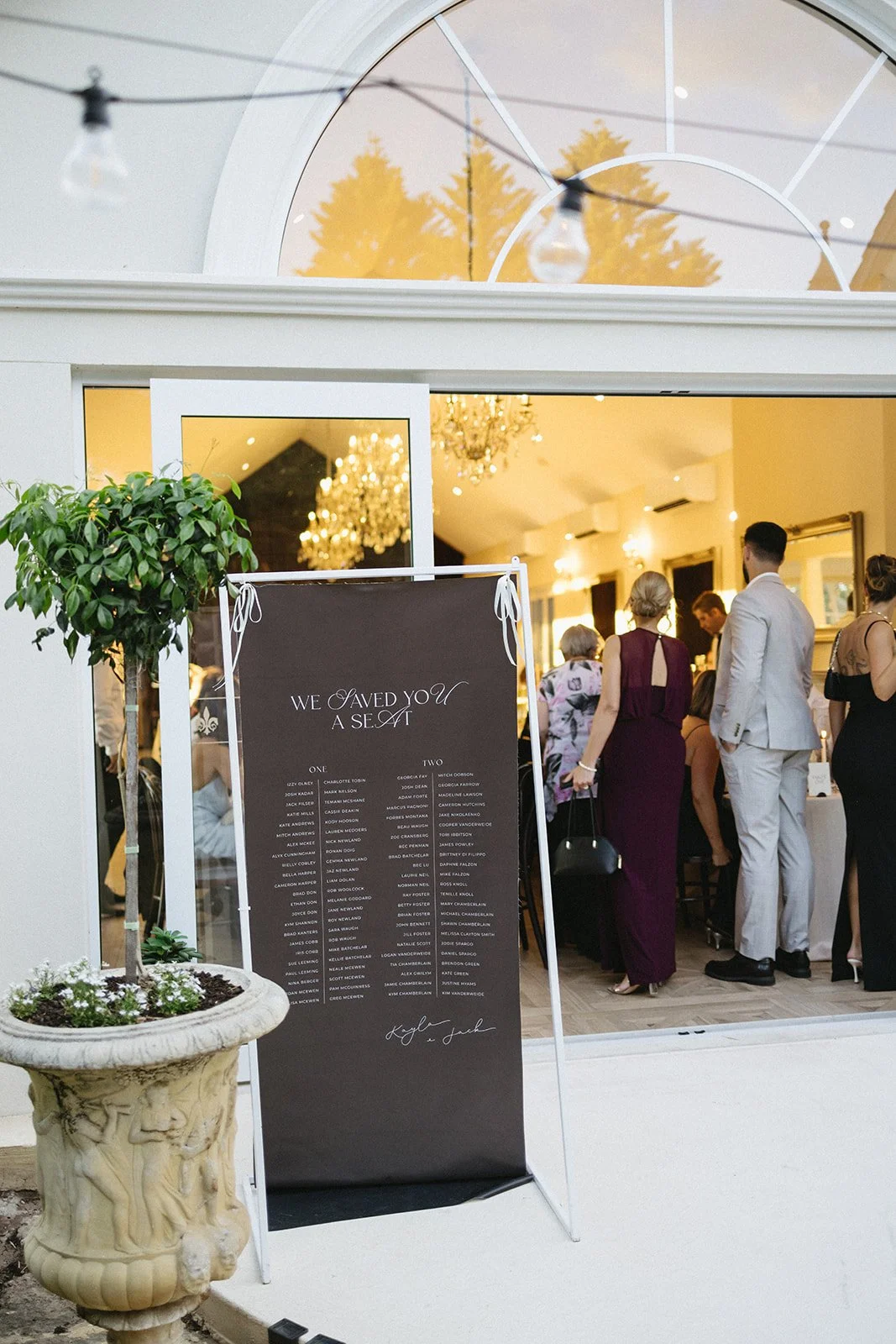 Guests at a wedding reception inside a well-lit hall, visible through an open door, with elegant chandeliers hanging from the ceiling. A large sign outside displays the guest list, with a potted plant nearby.
