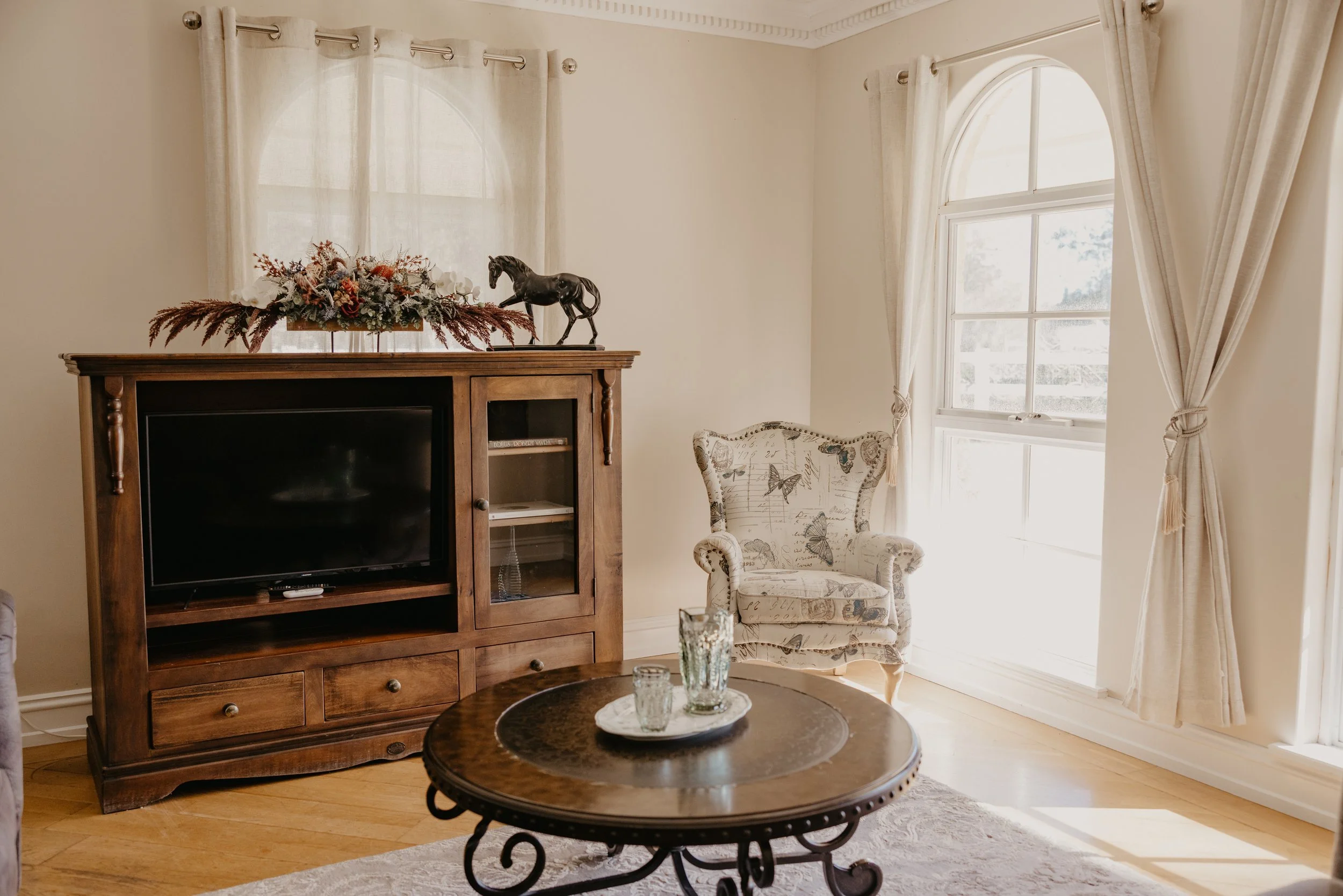 Living room with cream-colored walls, cream curtains, a wooden TV stand with a flat-screen TV, a decorative horse figurine, a floral arrangement on top, an upholstered armchair with butterfly and script print, and a round wooden coffee table with gla