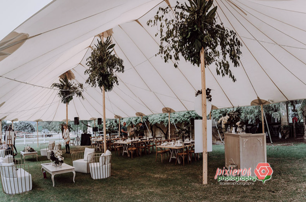 Outdoor event setup under a large white tent with decorated tables and seating, floral arrangements, and greenery.