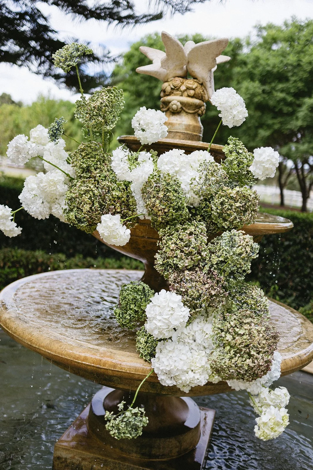 A multi-tiered water fountain decorated with white and green hydrangea flowers, set outdoors under a cloudy sky with trees in the background.