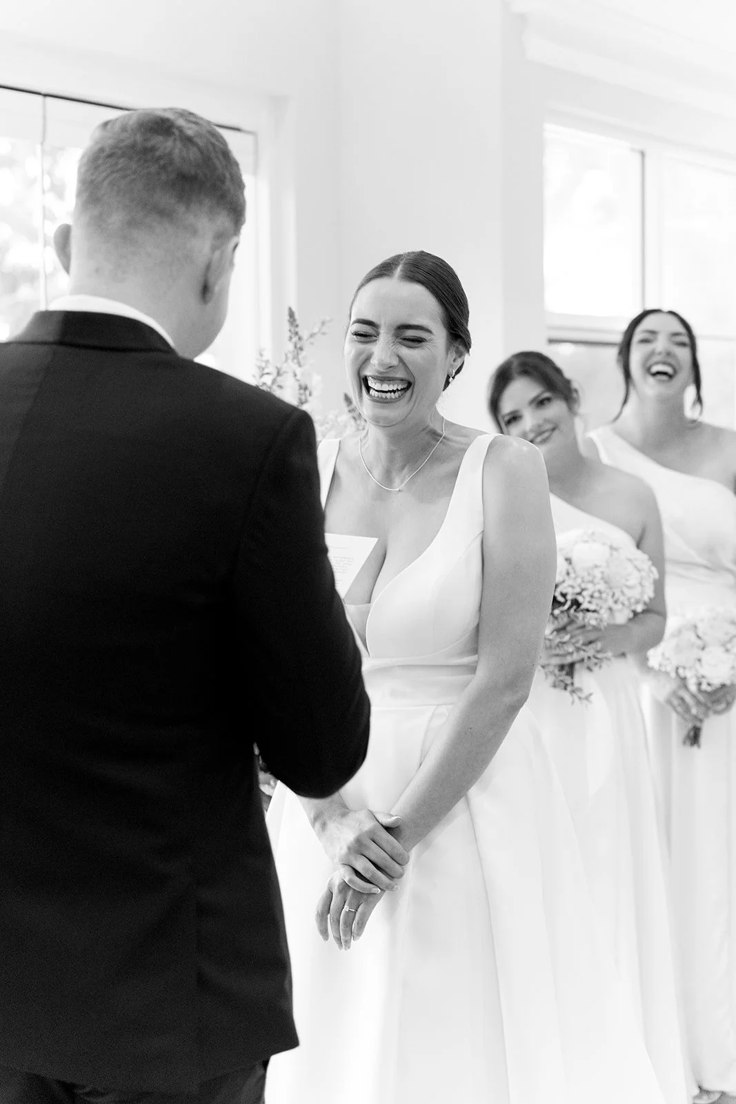 A bride laughing during her wedding ceremony, with her hand on her stomach, wearing a white wedding dress, and bridesmaids holding bouquets in the background.