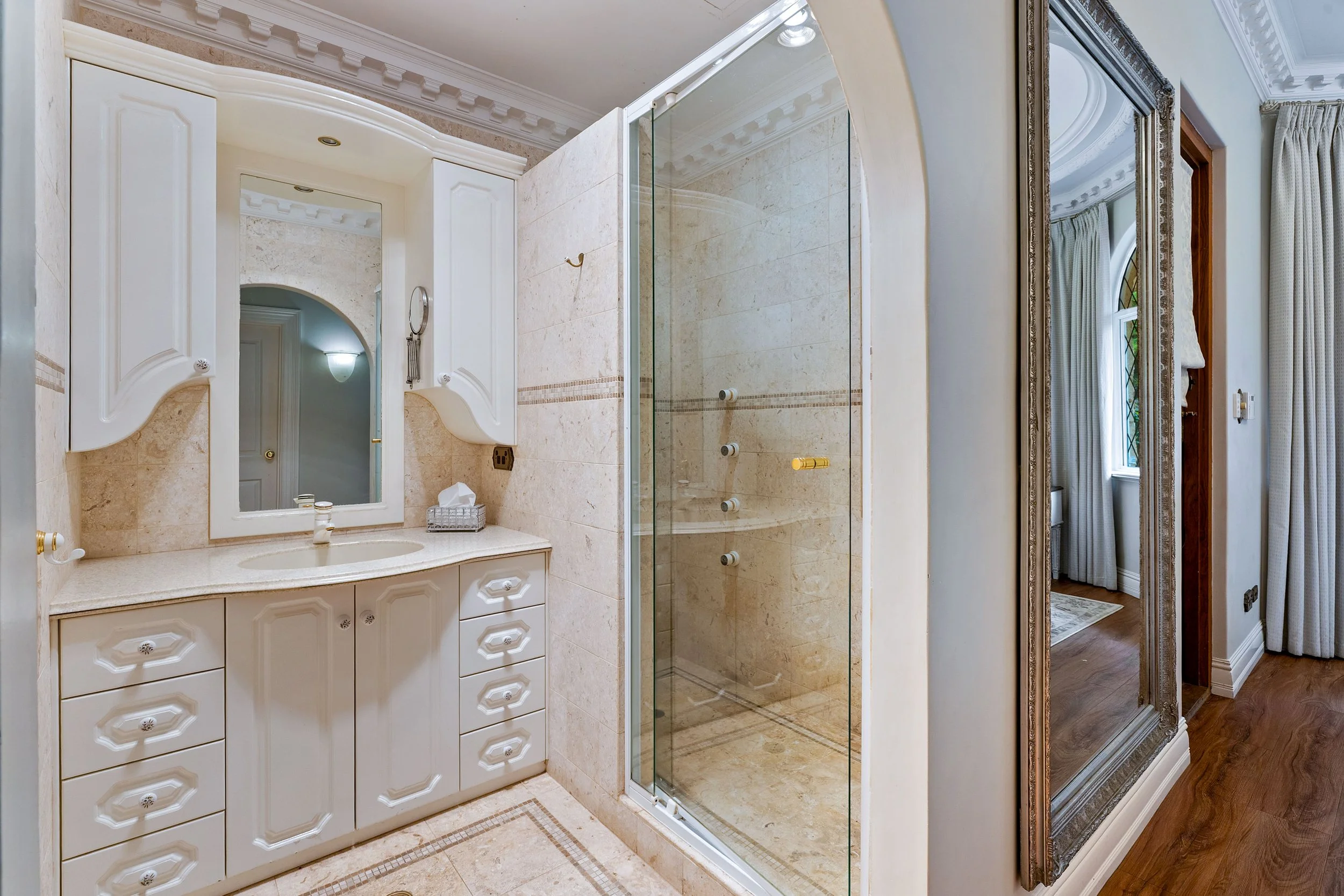 A bathroom featuring a white vanity with multiple drawers, a large mirror, a glass-enclosed shower with beige tiles and decorative border, and a mirror with an ornate frame reflecting a window with curtains.