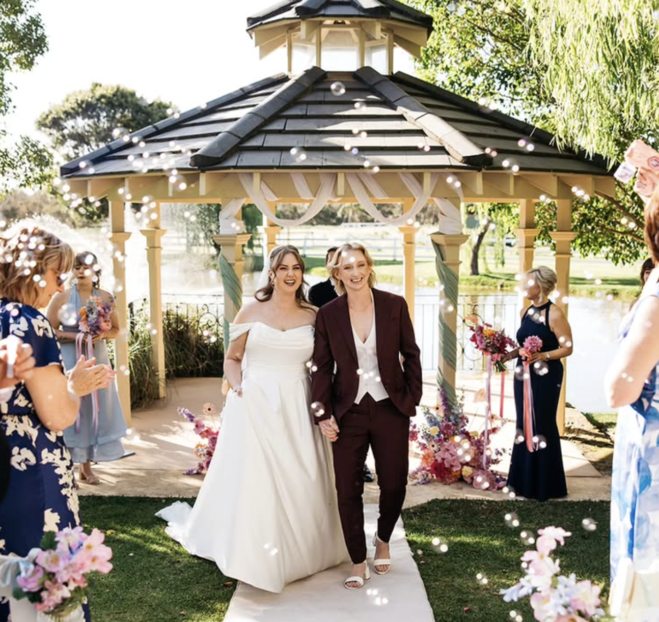 Two women, one in a wedding dress and the other in a suit, holding hands and walking down an outdoor aisle at a wedding ceremony. Guests are clapping and celebrating under a gazebo decorated with flowers and ribbons.