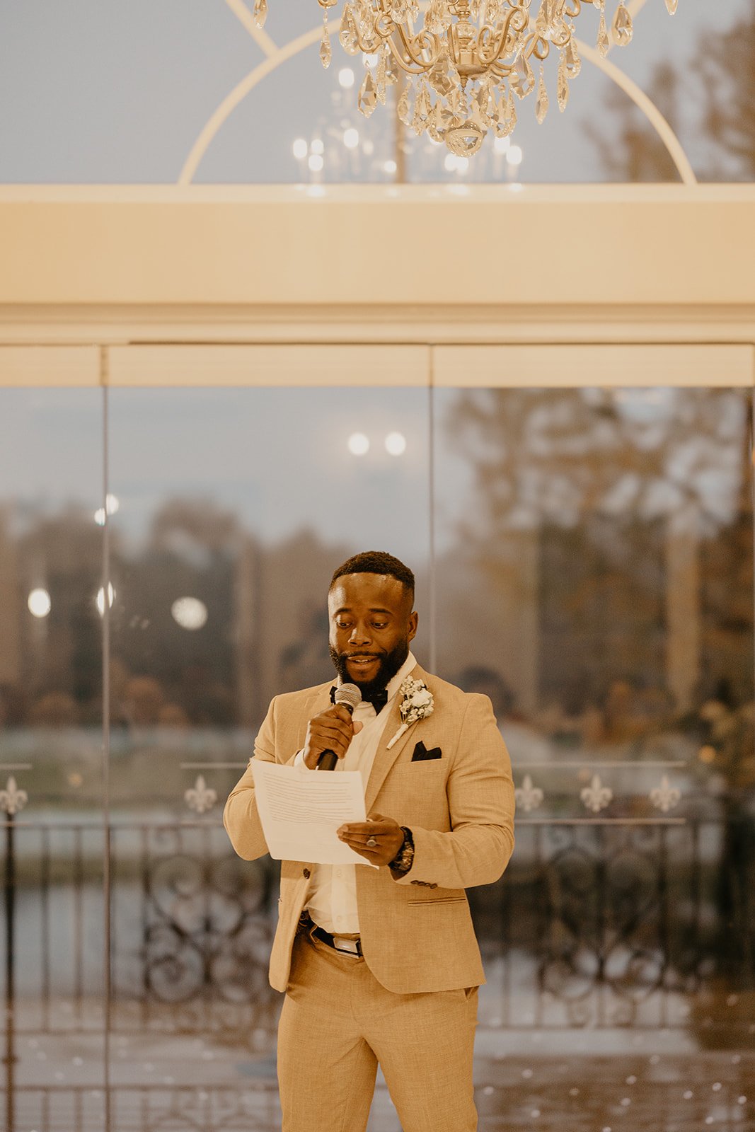 A man in a beige suit giving a speech at a wedding, holding a microphone and reading from a paper, with a chandelier above and large glass windows in the background.