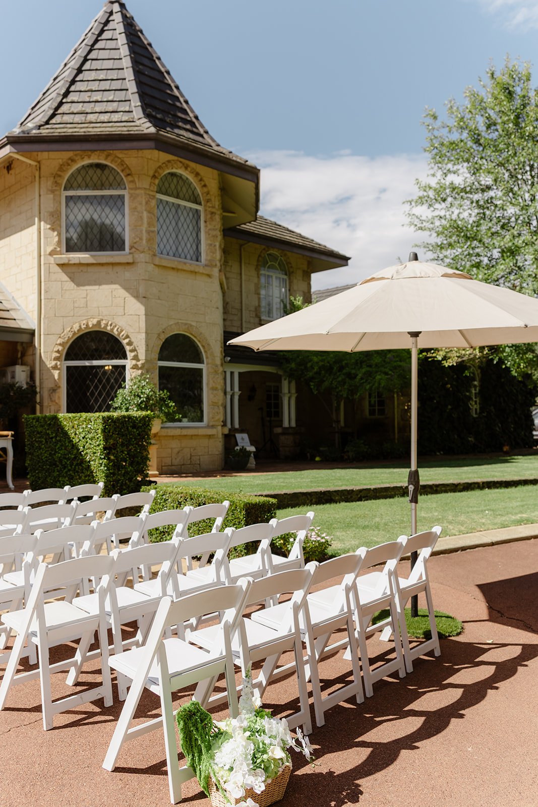 A wedding setup outdoors with white chairs arranged in rows on a brick pathway, a large white patio umbrella, and a large stone house with arched windows in the background. There is a basket of white flowers and greenery on the ground in front of the