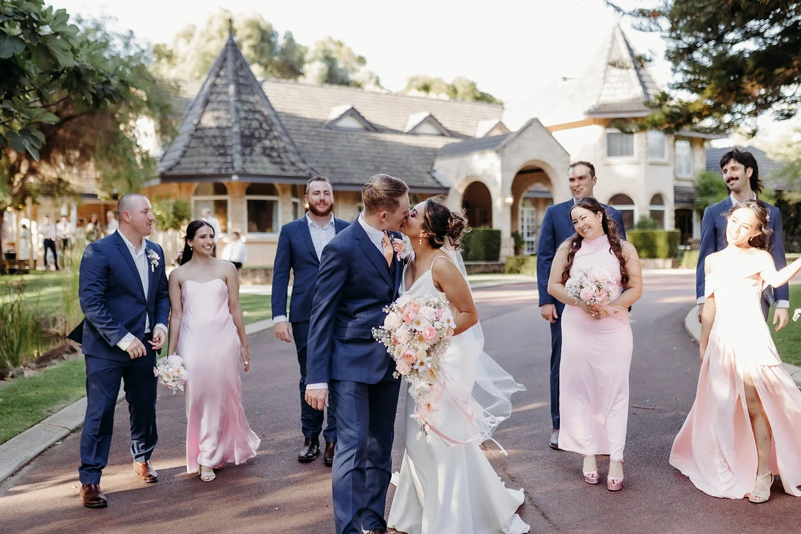 A wedding party celebrating outdoors in front of a large, elegant house. The bride and groom are kissing, with the bride holding a bouquet of pink and white flowers. Bridesmaids in pink dresses and groomsmen in blue suits are smiling and walking arou