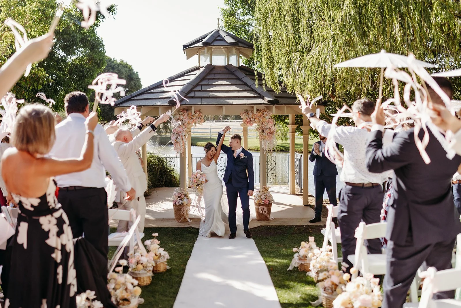A newlywed couple walking down the aisle at their outdoor wedding ceremony, surrounded by guests celebrating and waving ribbons, with floral decorations and a gazebo in the background.