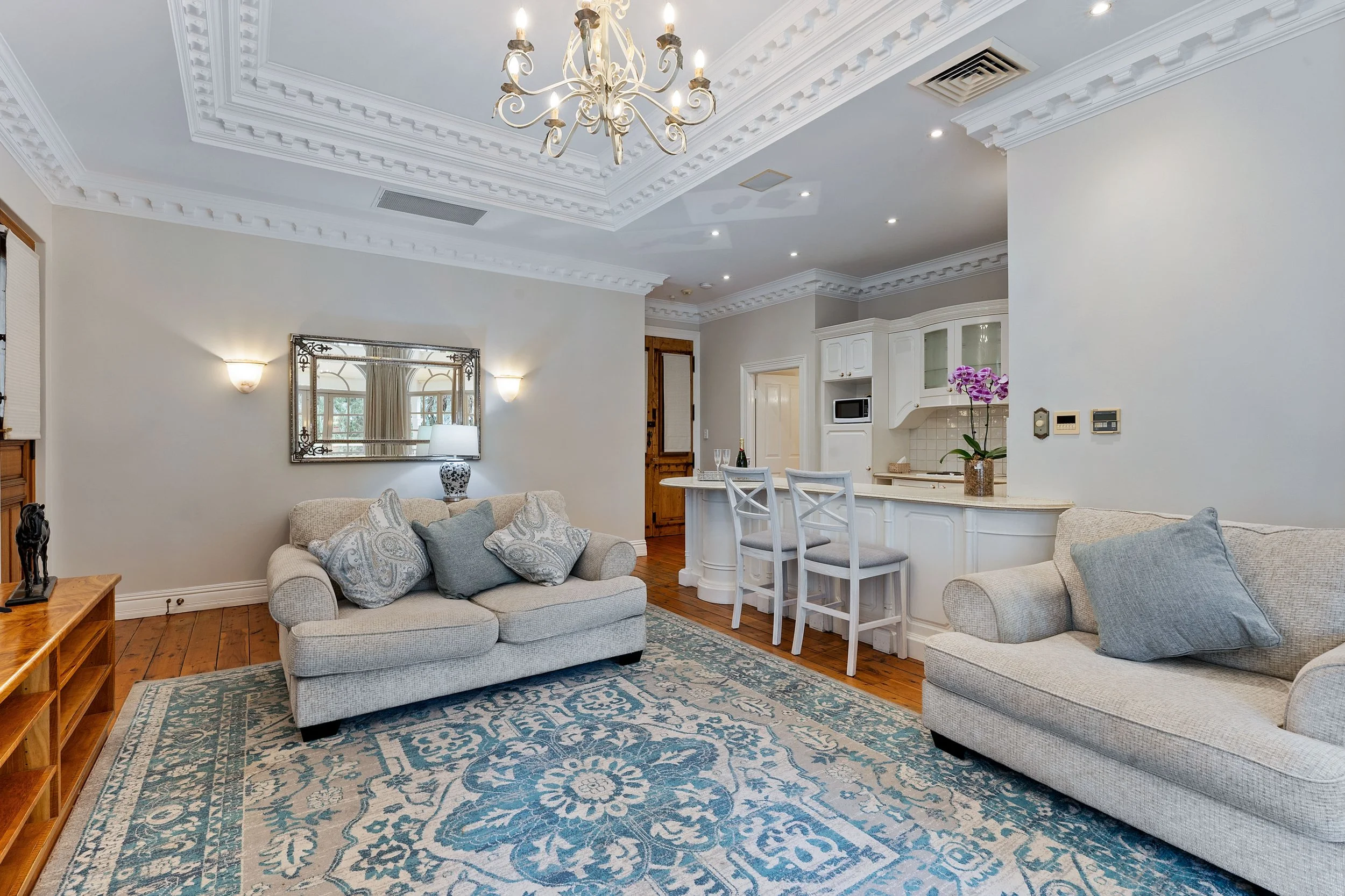 Living room with light-colored sofas, a large patterned rug, a coffee table, wall sconces, a dining area with chairs, a chandelier, and a kitchen area with white cabinets and a purple orchid on the counter.