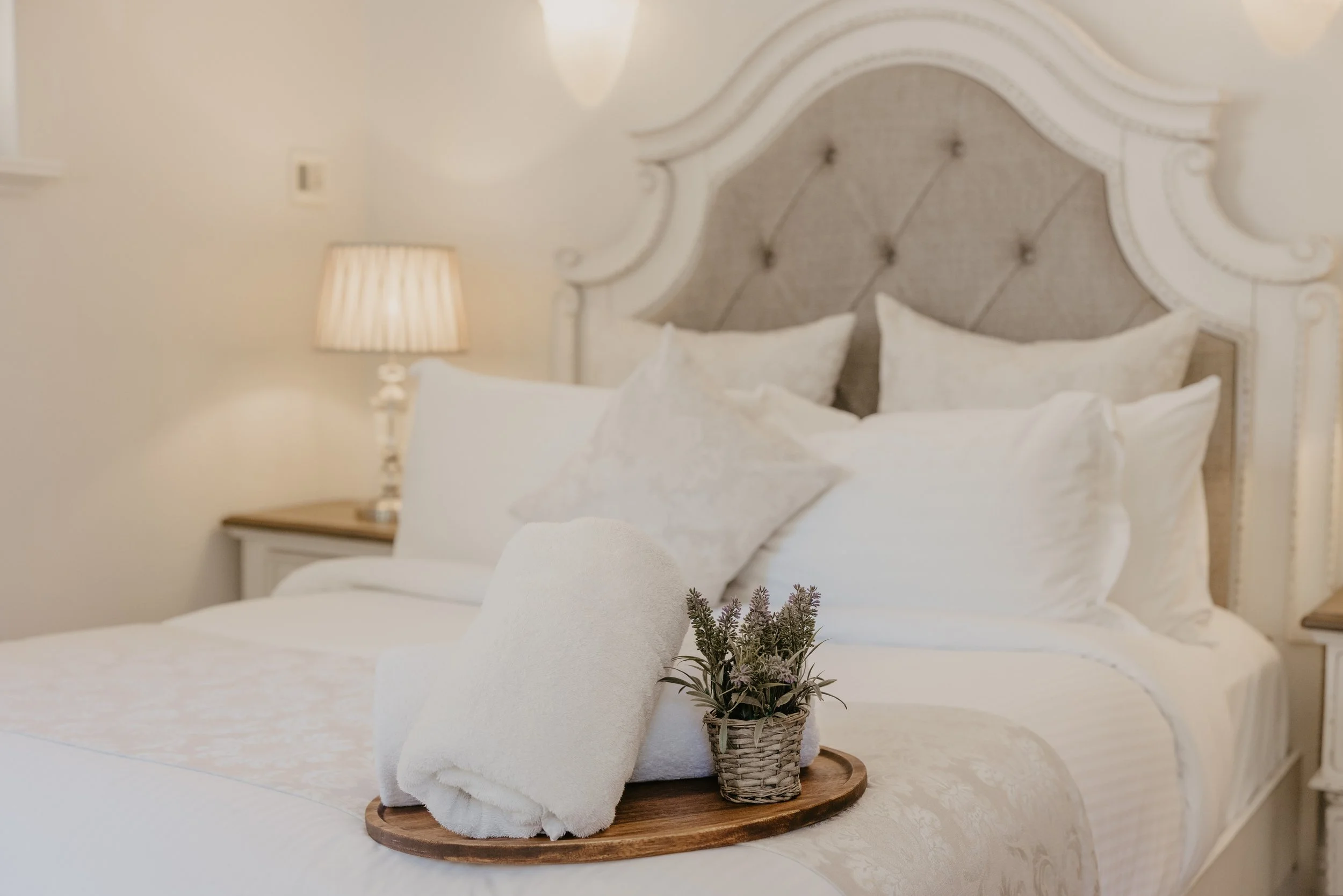 A decorative tray on a bed with white towels, a small potted plant, and bedding with pillows and a headboard in the background.