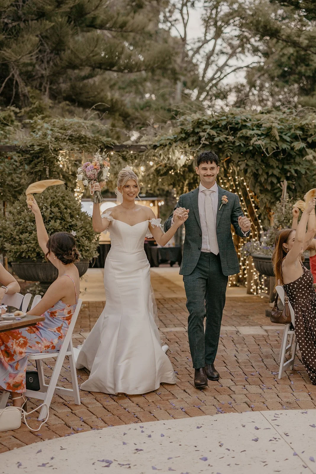 A newly married couple holding hands and celebrating at their outdoor wedding reception during daylight, with guests seated around and string lights in the background.