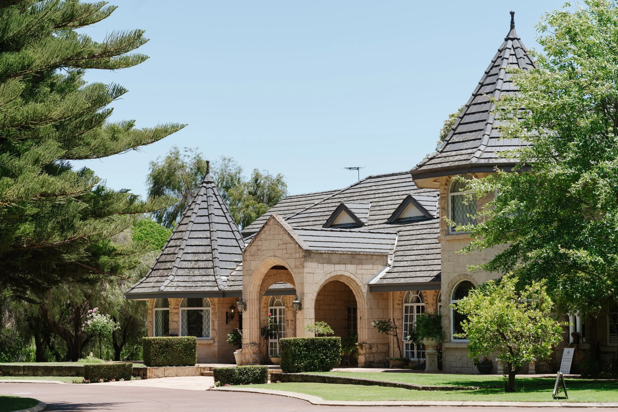 A stone house with a turret and arched entryway, surrounded by trees and manicured bushes, under a clear blue sky.