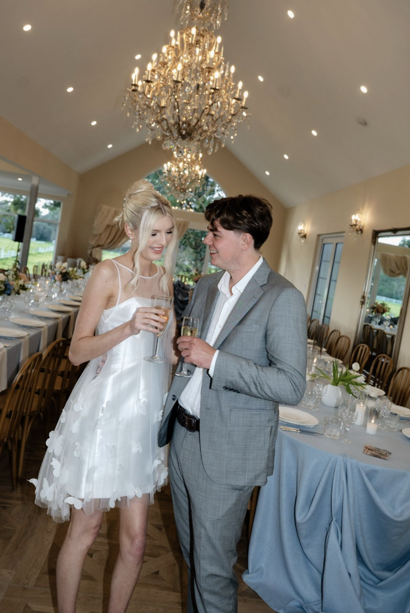 A young woman and man dressed in formal attire, holding champagne glasses, standing in a decorated event hall with chandeliers and a long dining table.