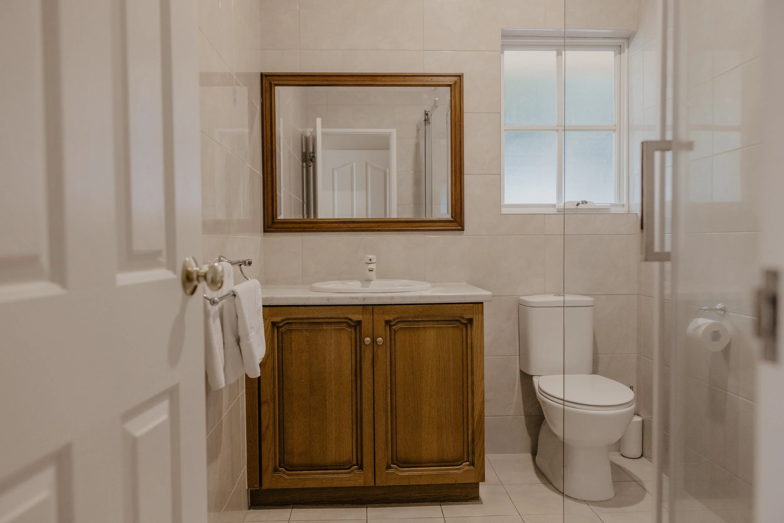 A bathroom with a wooden cabinet, a mirror, a sink, a toilet, a window, and tiled walls and floor.