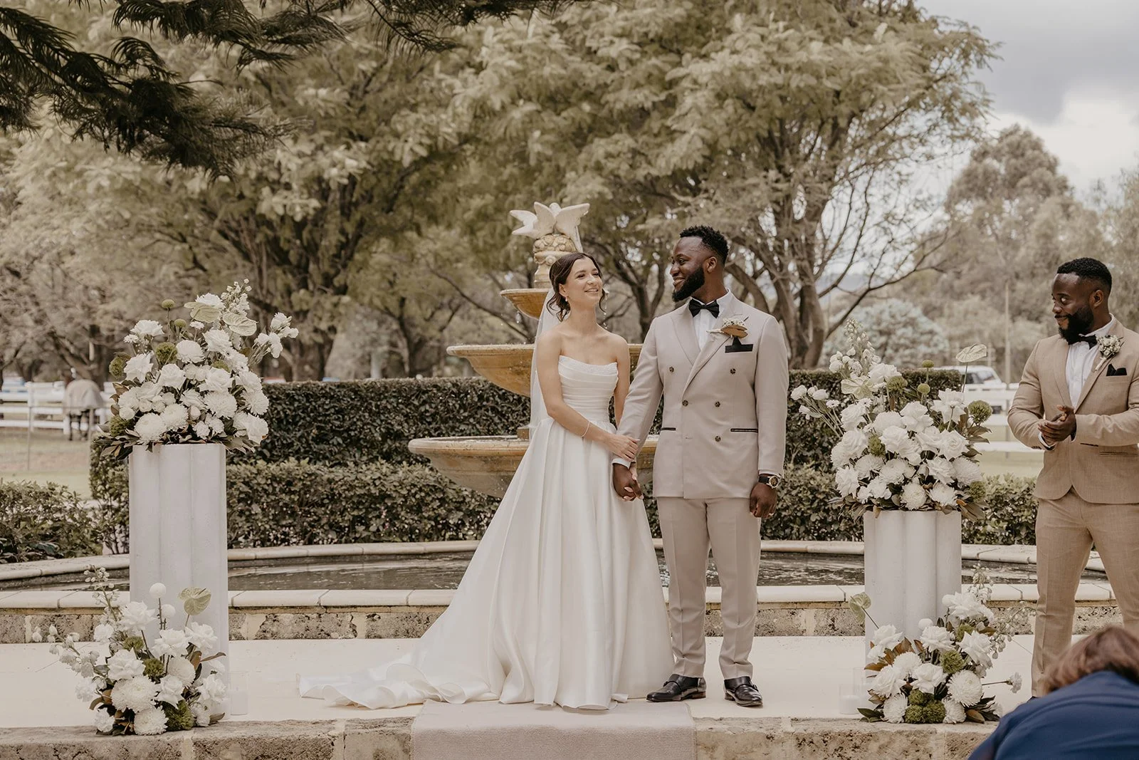 Bride and groom holding hands during their wedding ceremony outdoors, with officiant and decorative floral arrangements nearby, in front of a fountain with trees in the background.