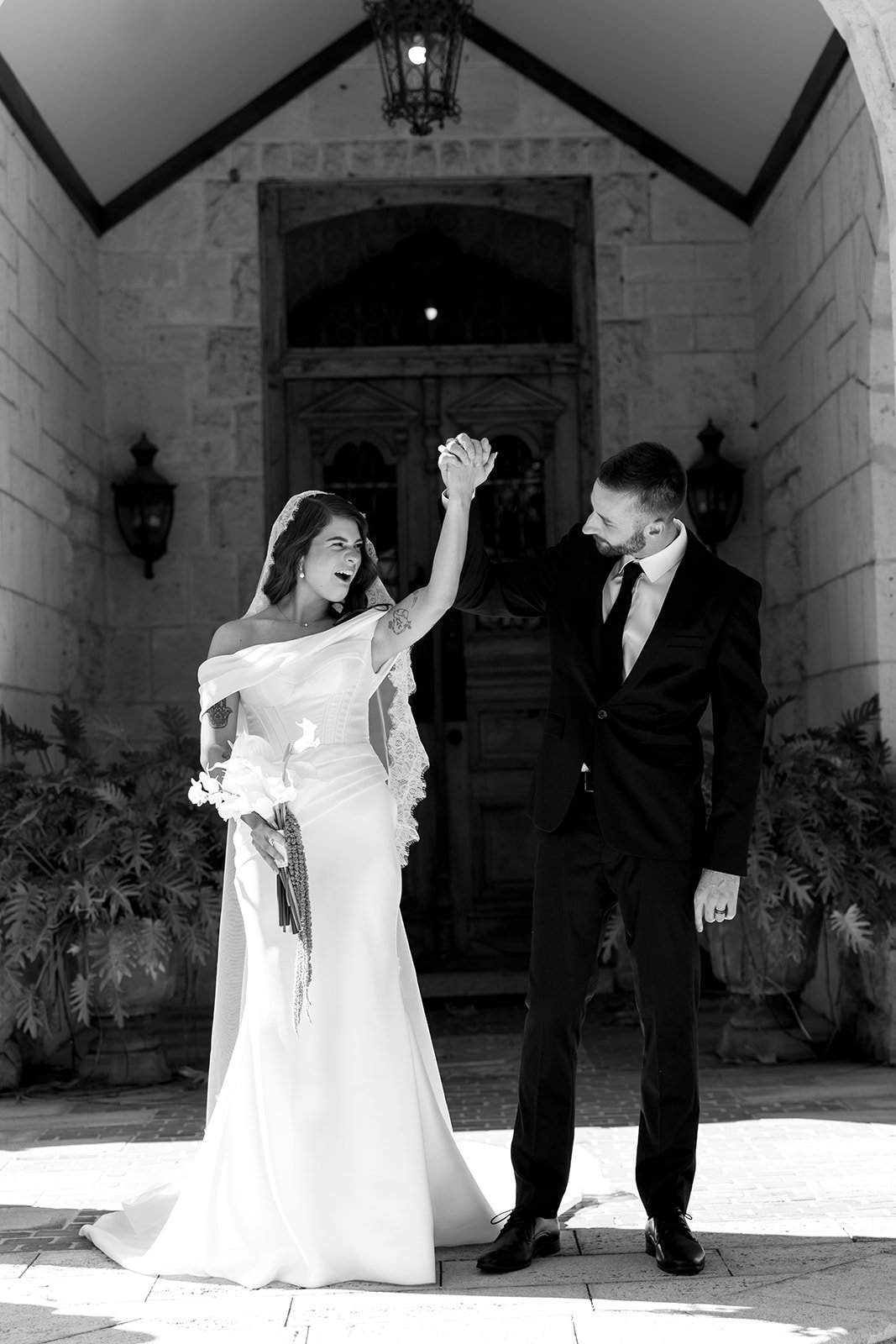 Black and white photo of a bride and groom celebrating their wedding, holding hands in the air, standing outside a building with stone walls and a wooden door.