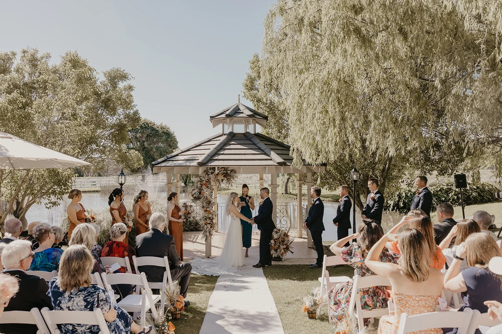 Outdoor wedding ceremony at  Brookleigh Estate with bride and groom holding hands under a gazebo, surrounded by bridesmaids and groomsmen, guests seated on white chairs, scenic water and trees in the background.