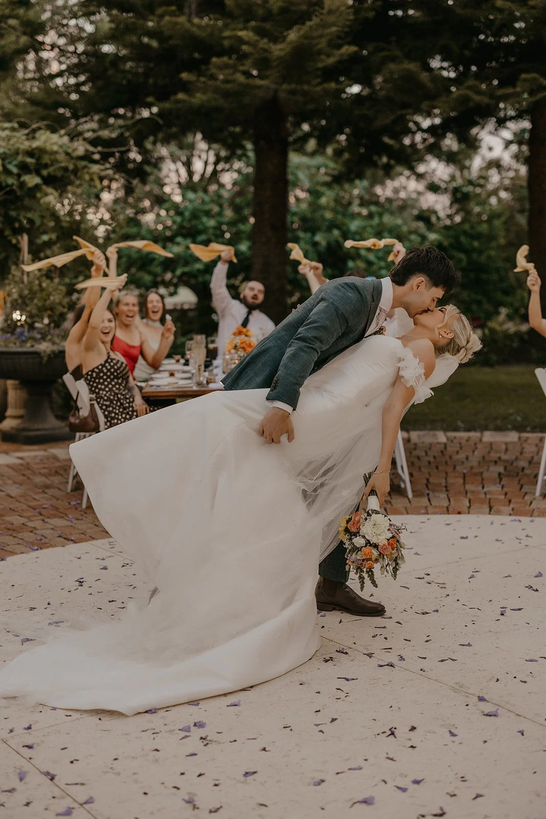A newlywed couple kisses as the groom dips the bride. Guests cheer and throw ribbons in the background at an outdoor wedding reception.