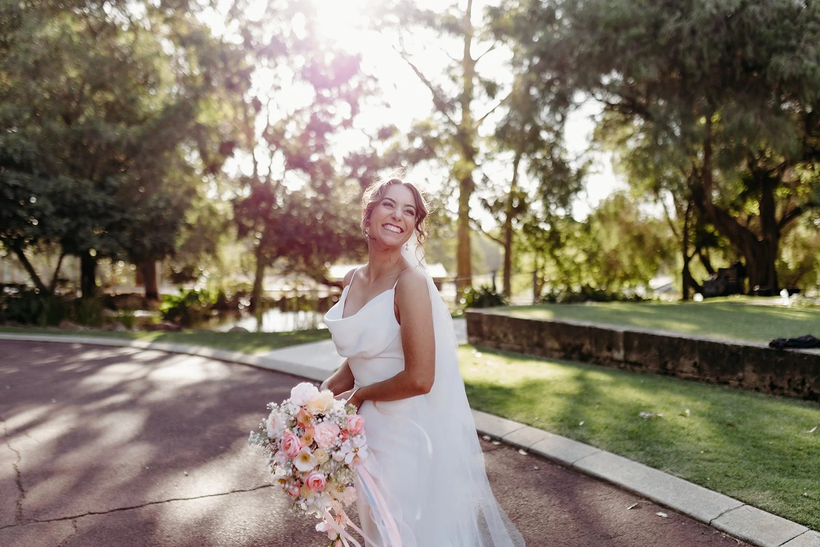 A smiling bride in a white wedding dress holding a bouquet of pink and white flowers outdoors in a park with trees and sunlight in the background.