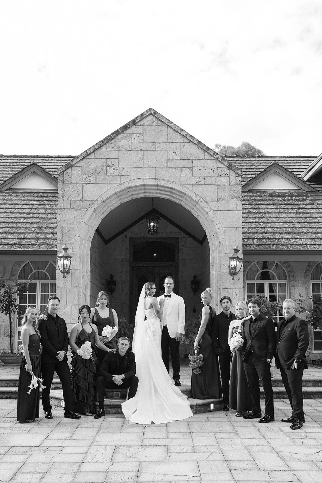 Black and white photo of a wedding party standing on front steps of a stone building with an arched entrance. The bride and groom are in the center, with the bride in a long white dress and veil, and the groom in a white tuxedo. Several bridesmaids i