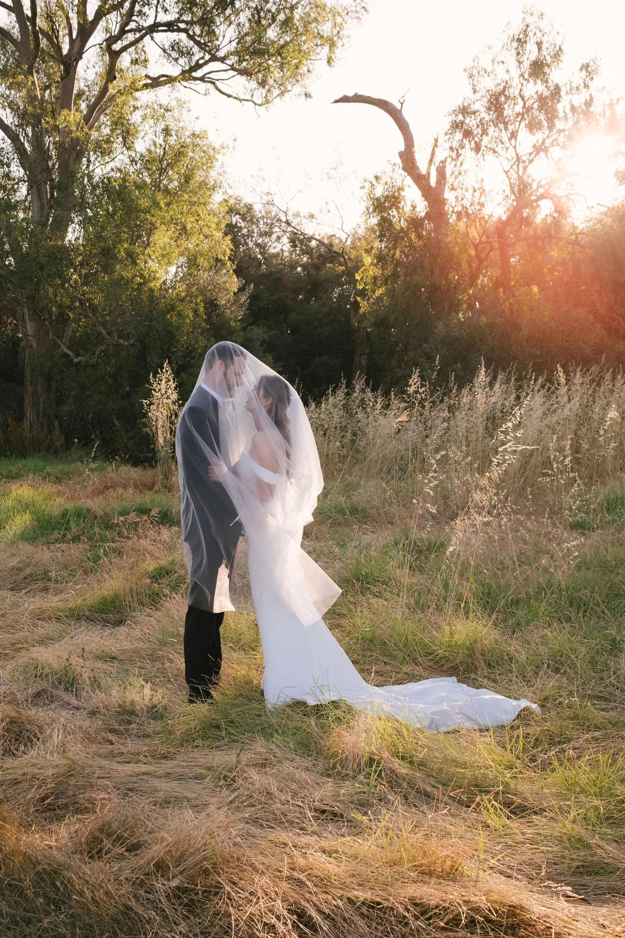 A bride and groom embrace under a veil in a grassy field at sunset, surrounded by trees.