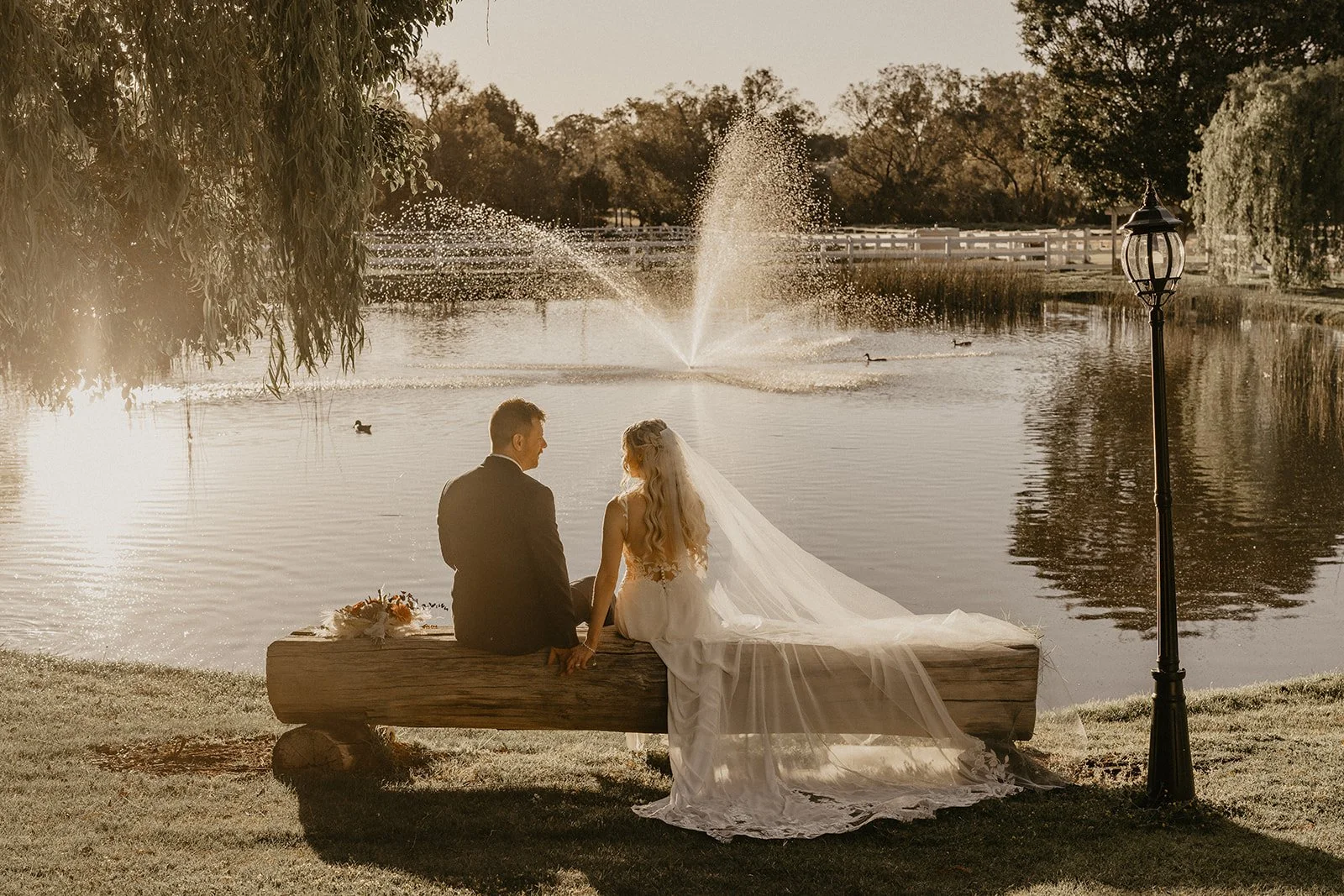 A bride and groom sitting on a wooden bench by a pond, holding hands and facing each other, during sunset with a fountain spraying water in the background in the Swan Valley Perth at Brookleigh Estate