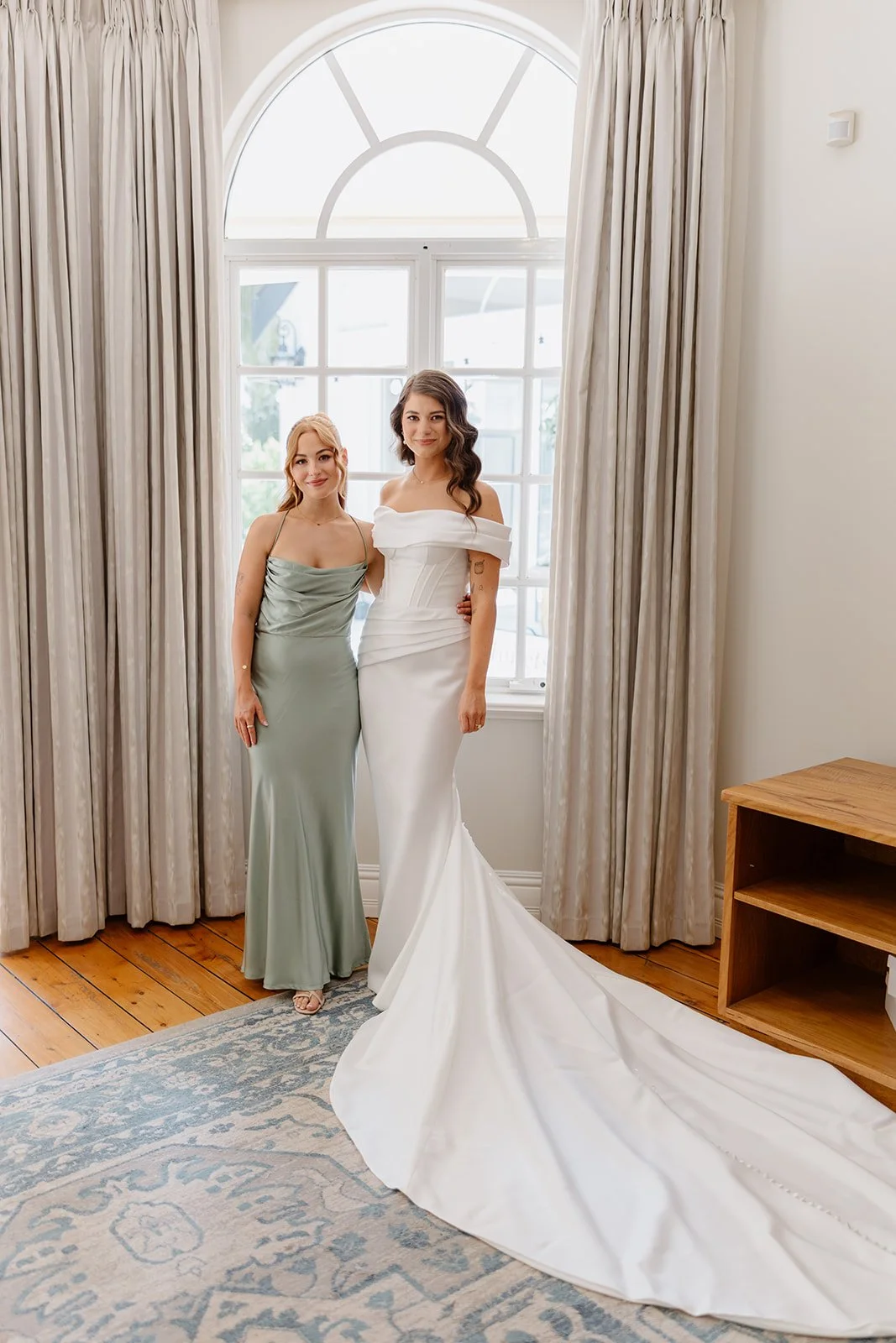 Two women standing side by side in front of a large window, one in a wedding dress and the other in a formal gown, inside a well-lit room with curtains and a patterned rug.