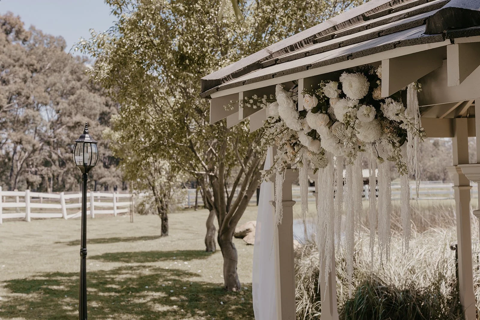 White floral arrangement decorated with white flowers and lace hanging from a pavilion at a rustic outdoor wedding venue.
