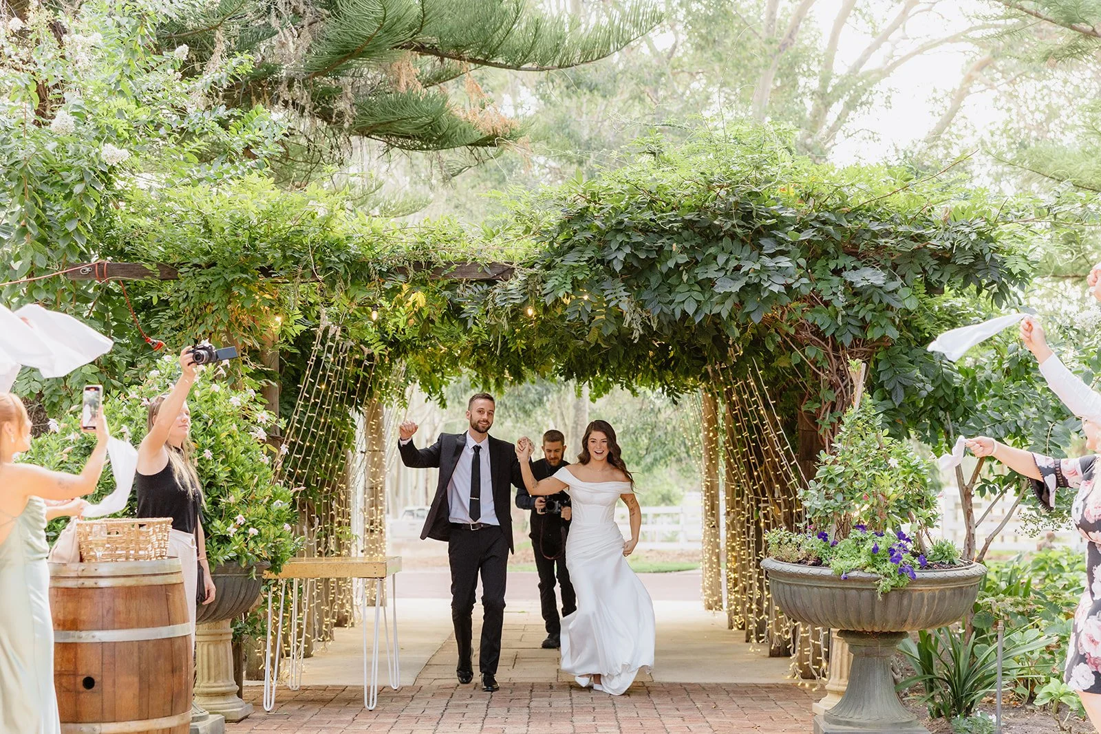 A newlywed couple walking through a decorated garden archway, smiling and celebrating, with guests surrounding and taking photos.