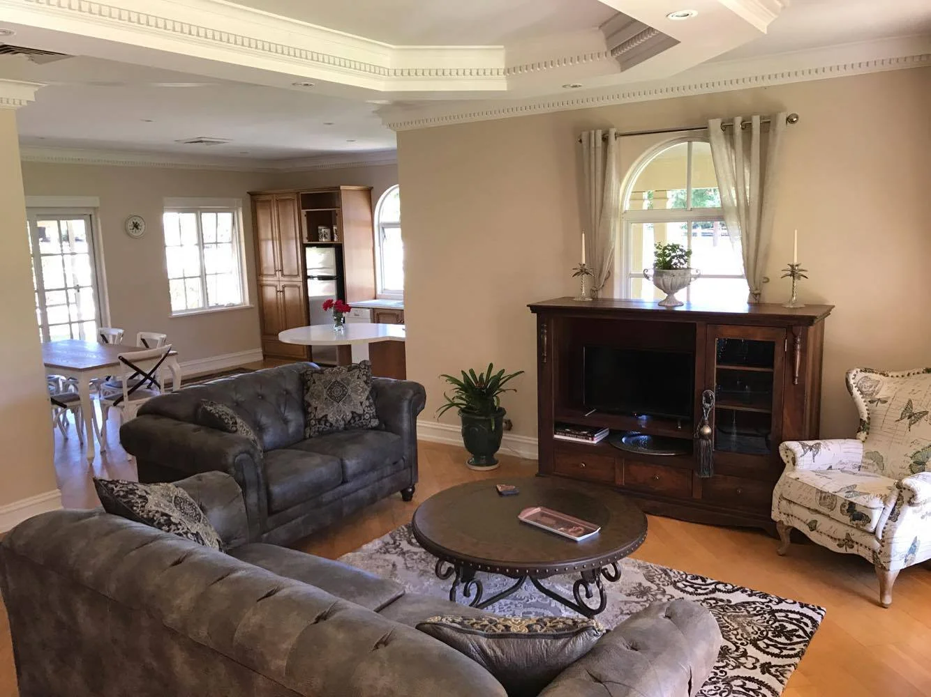 Living room with two brown sofas, a round wooden coffee table, a vintage armchair with floral upholstery, a wooden TV cabinet, and a plant by the window. The room is decorated with curtains, candles, and a rug.