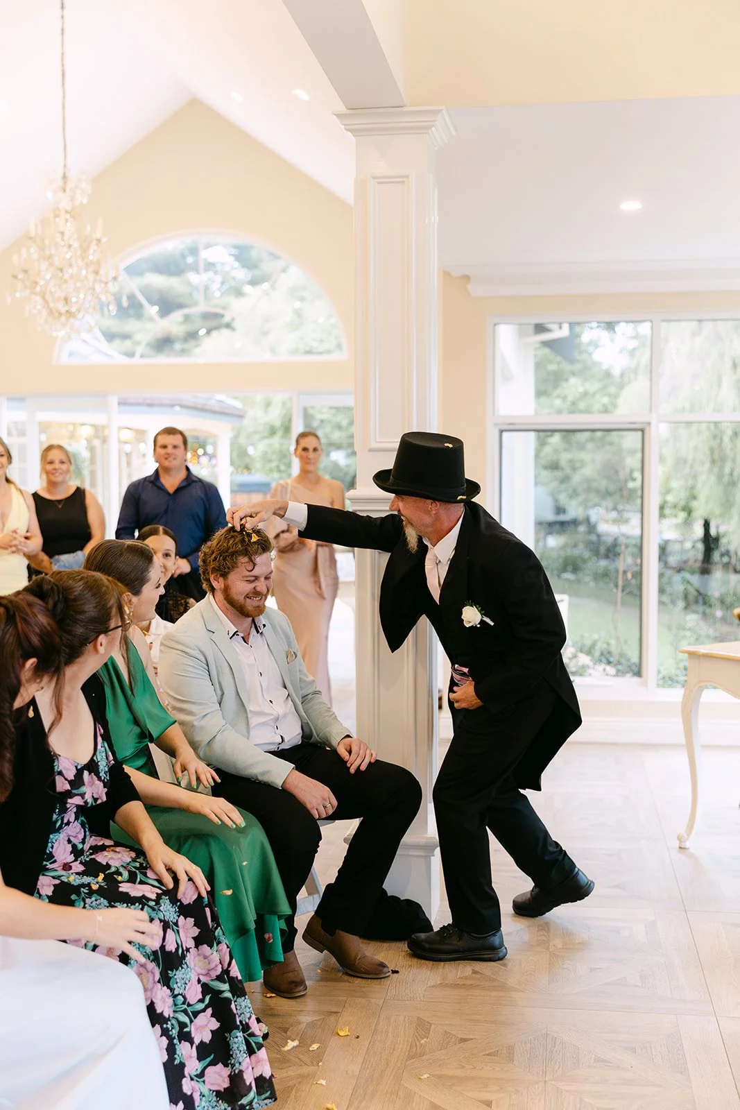 A man in a black suit and top hat is performing a magic trick or entertaining guests at a wedding reception. He is bending down in front of a seated man with curly hair and a light-colored blazer, who is smiling. Several guests are seated and watchin