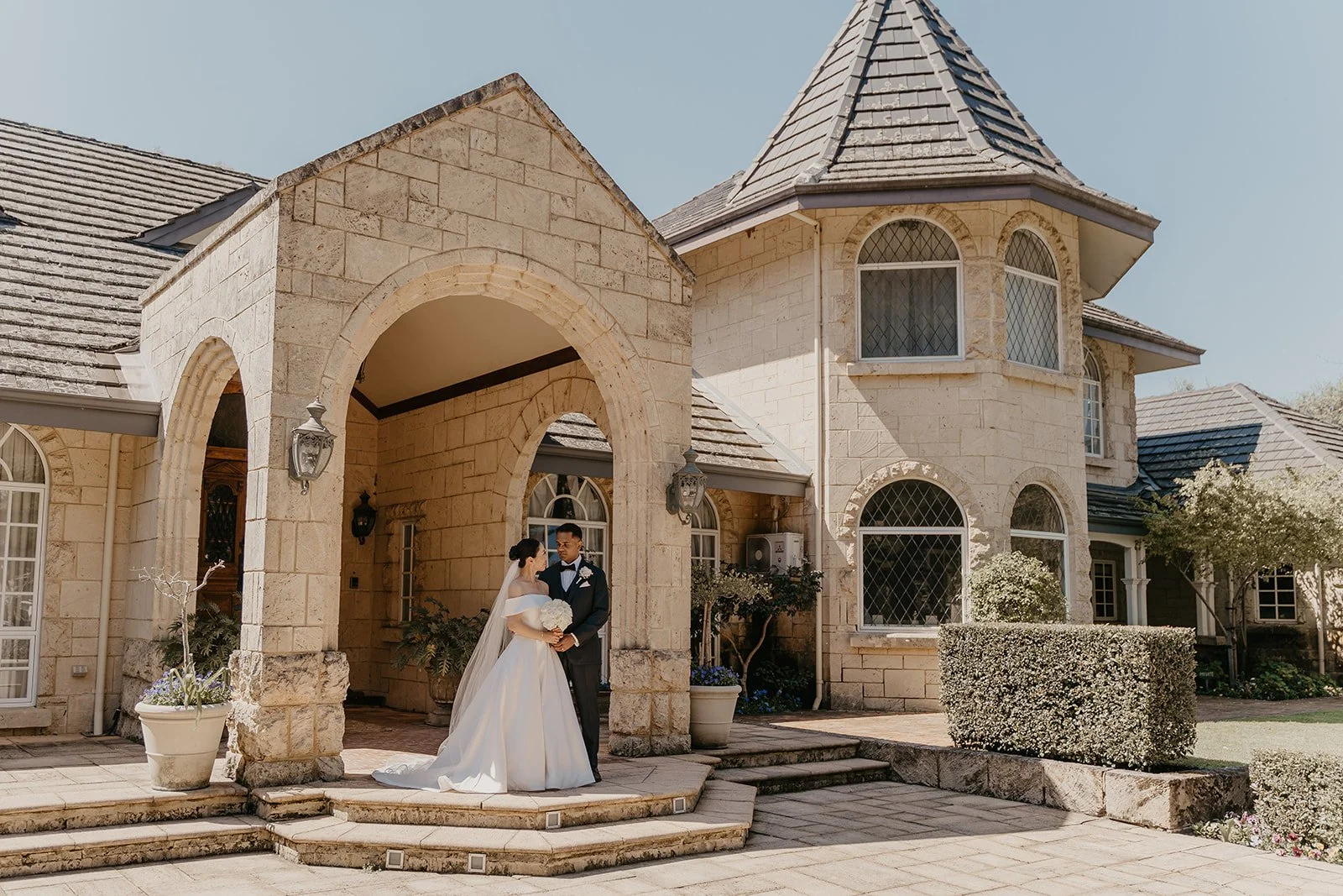 A bride and groom in wedding attire standing on steps outside a large stone house with arched windows and a turret, during daytime.
