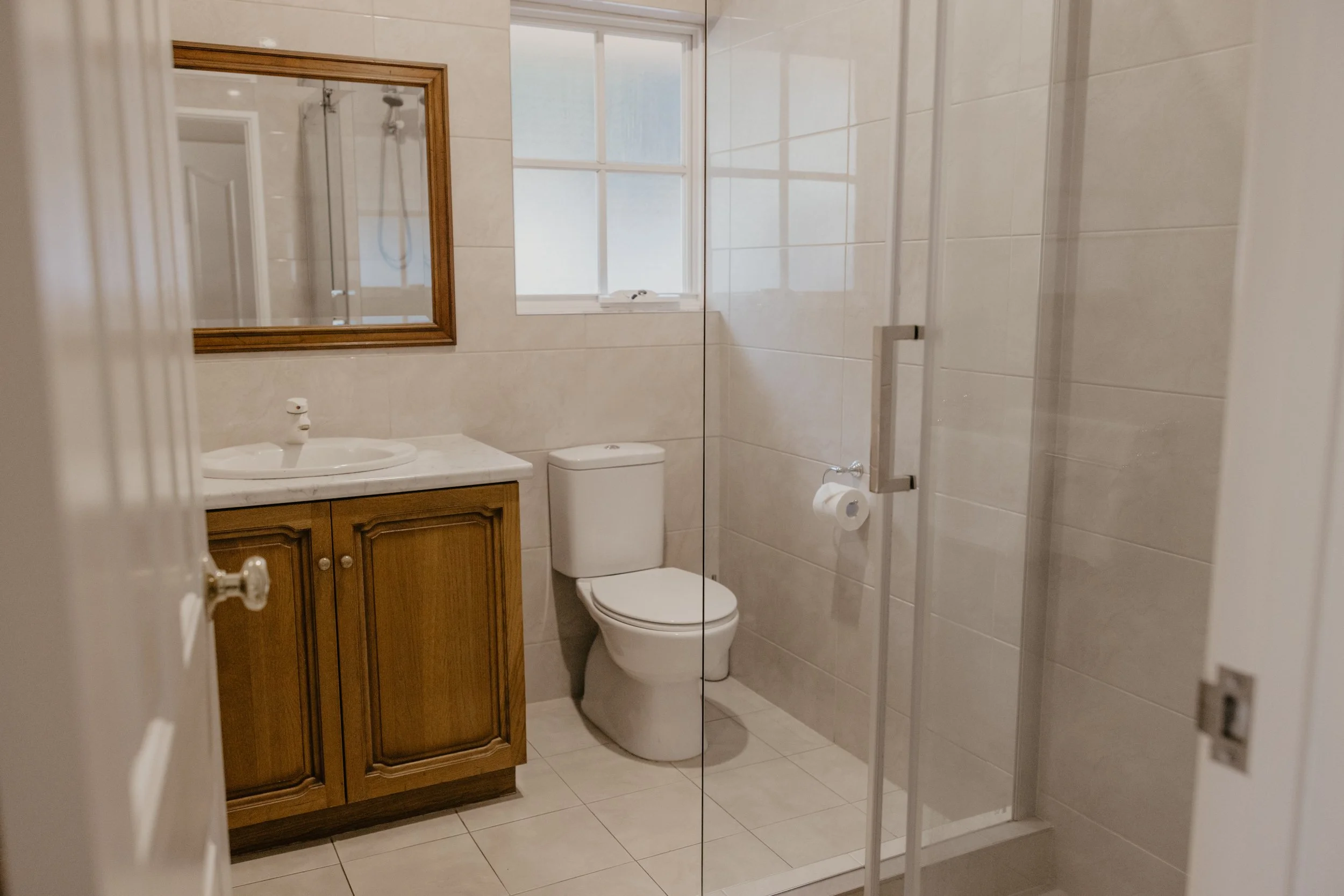 Bathroom with a wooden cabinet sink, a toilet, a mirror, and a shower with a glass door, beige tiled walls, and a window.
