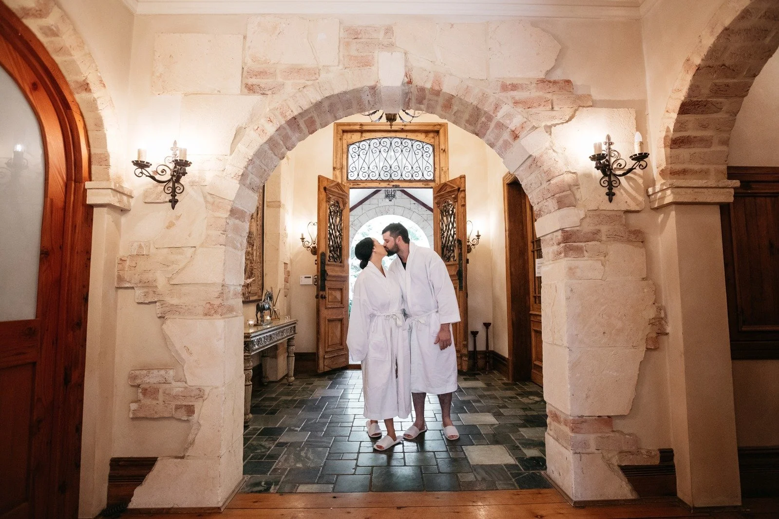 A couple in white robes sharing a kiss in a rustic, well-lit foyer with stone archways, wooden doors, and vintage wall sconces.