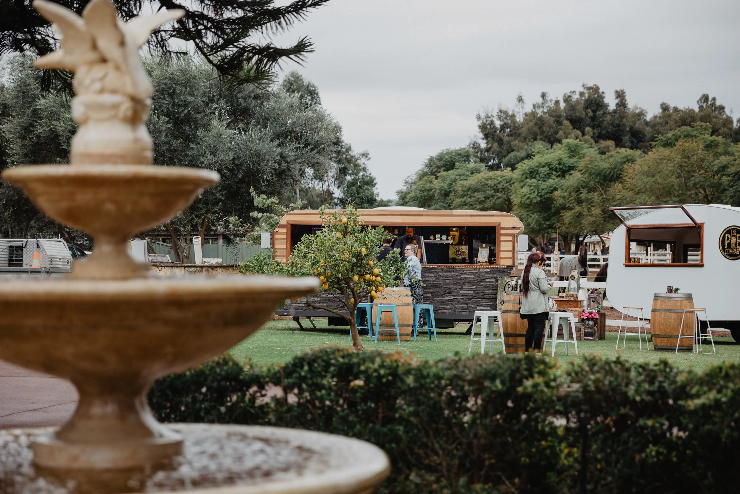 Outdoor scene with food trucks, people, and trees, viewed through a decorative stone fountain.