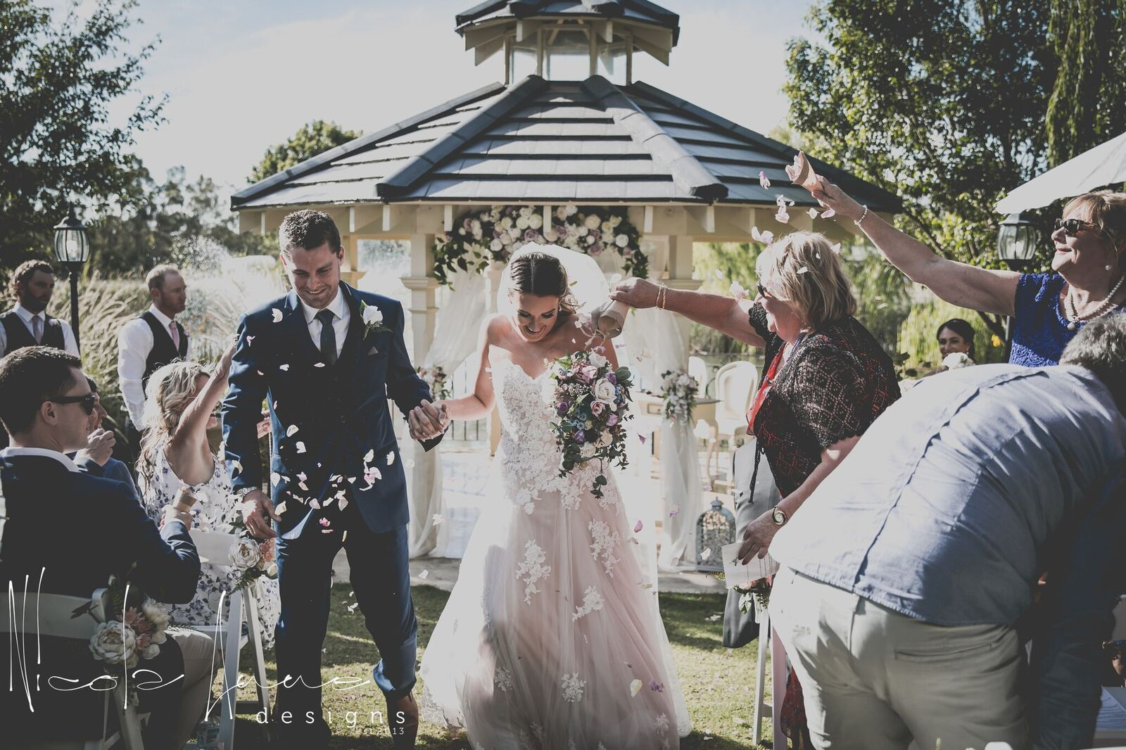 Bride and groom walking through confetti at outdoor wedding ceremony with guests celebrating and flowers in background.