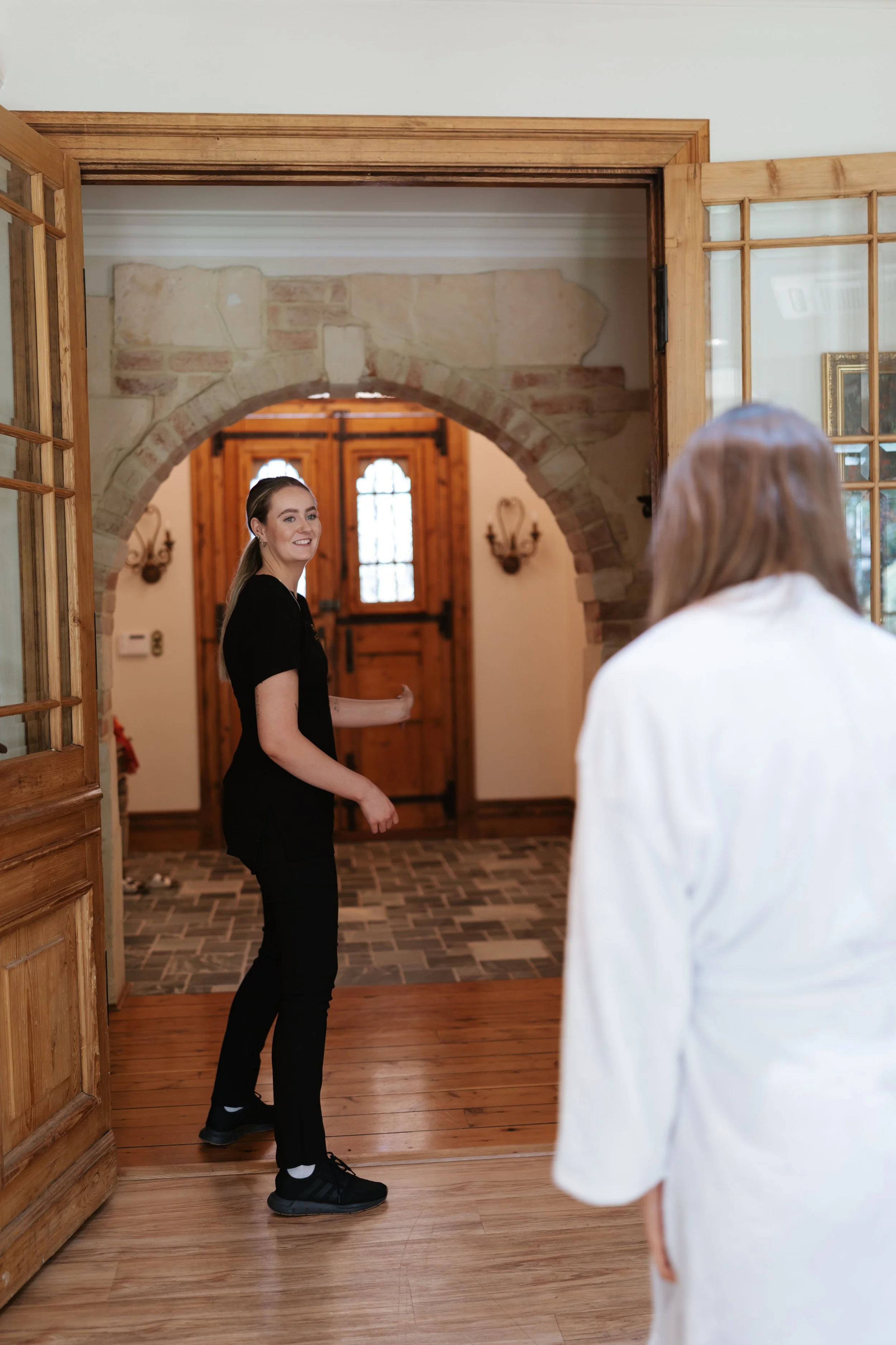 A young woman in a black uniform happily greets an older woman in a white coat at the entrance of a house with wooden and stone features.