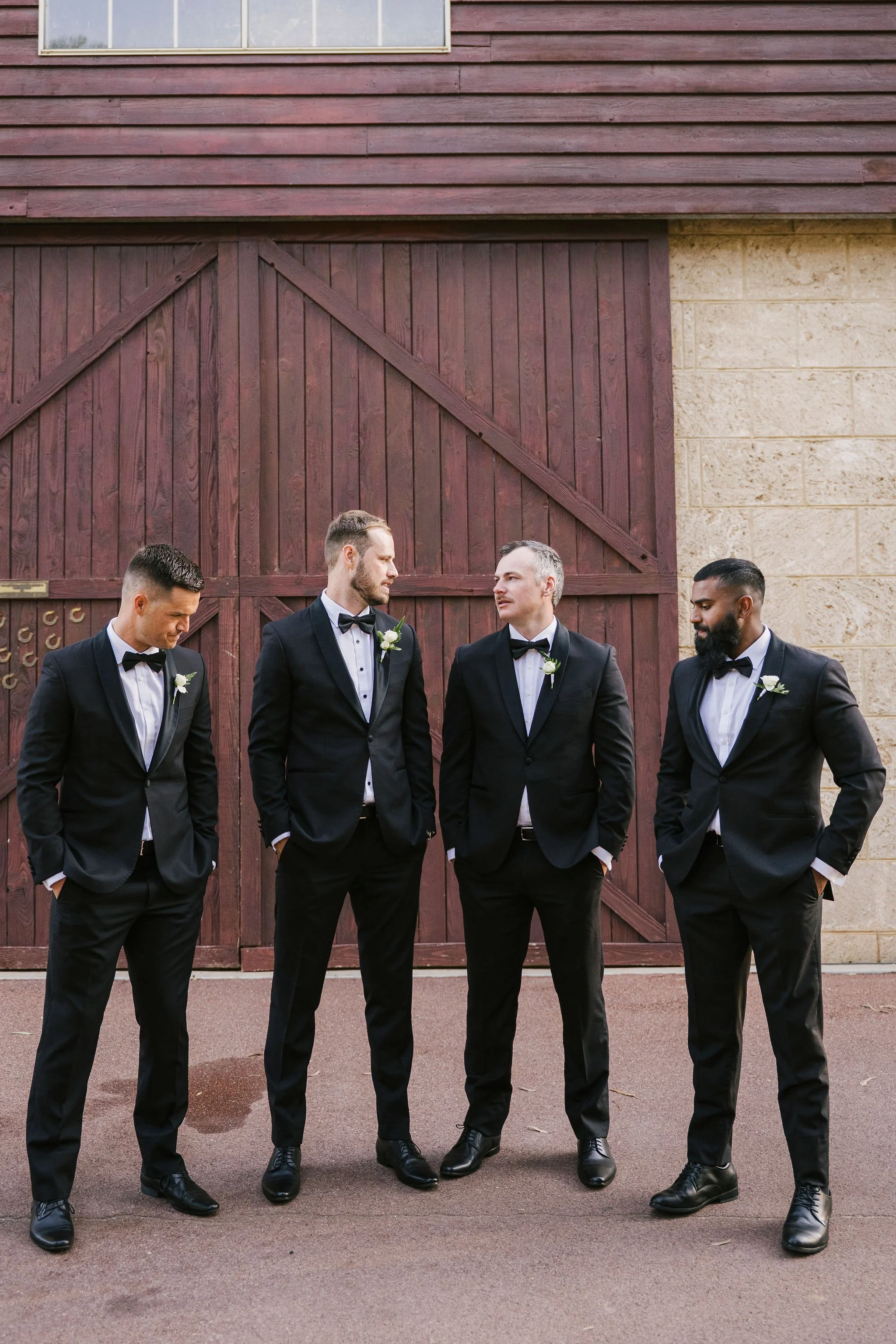 Four men in tuxedos standing in front of a wooden barn door, engaging in conversation.