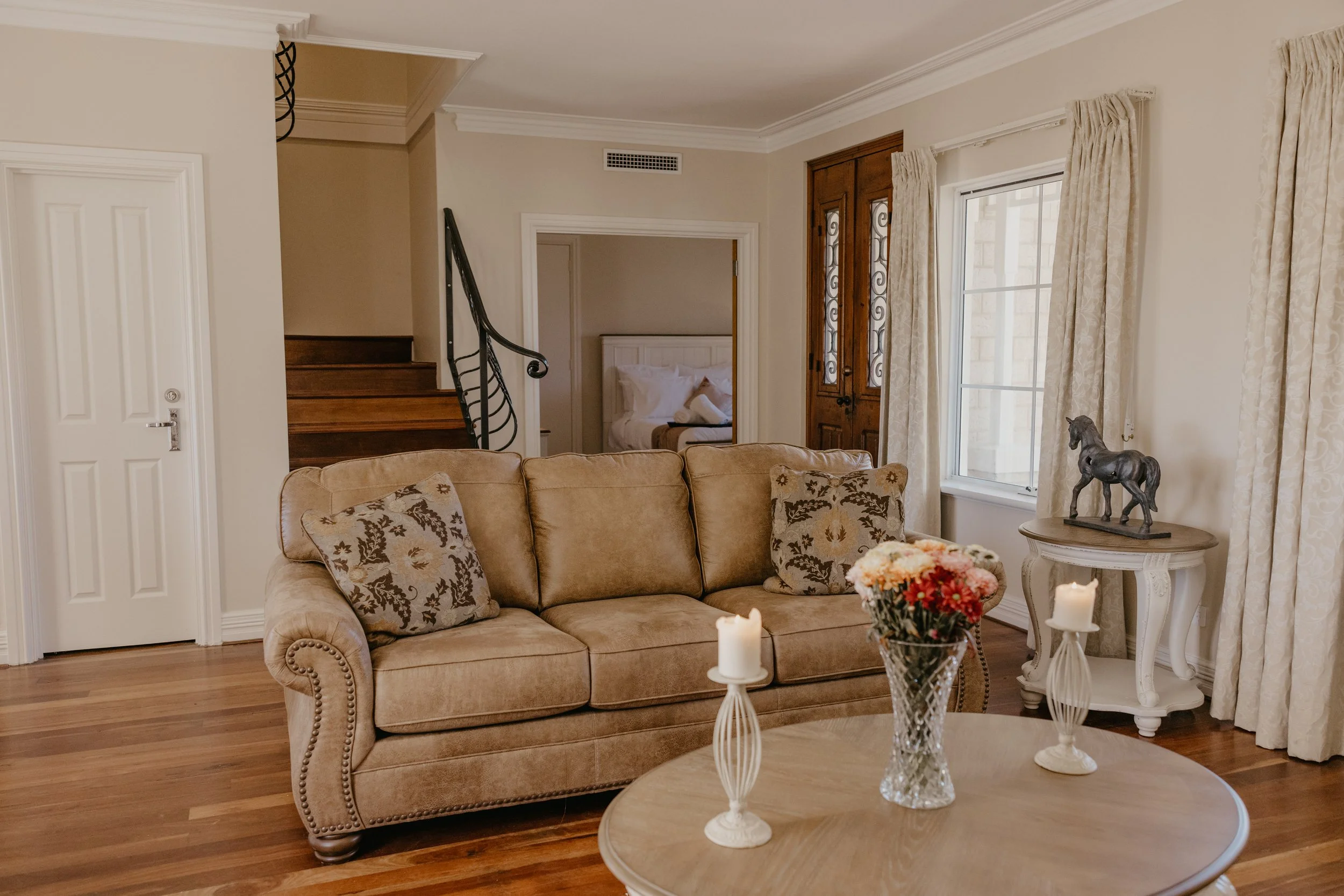 Living room with beige sofa, floral pillows, white side table with a horse figurine, curtains, window, wooden stairs, bedroom doorway, and candles on a coffee table.