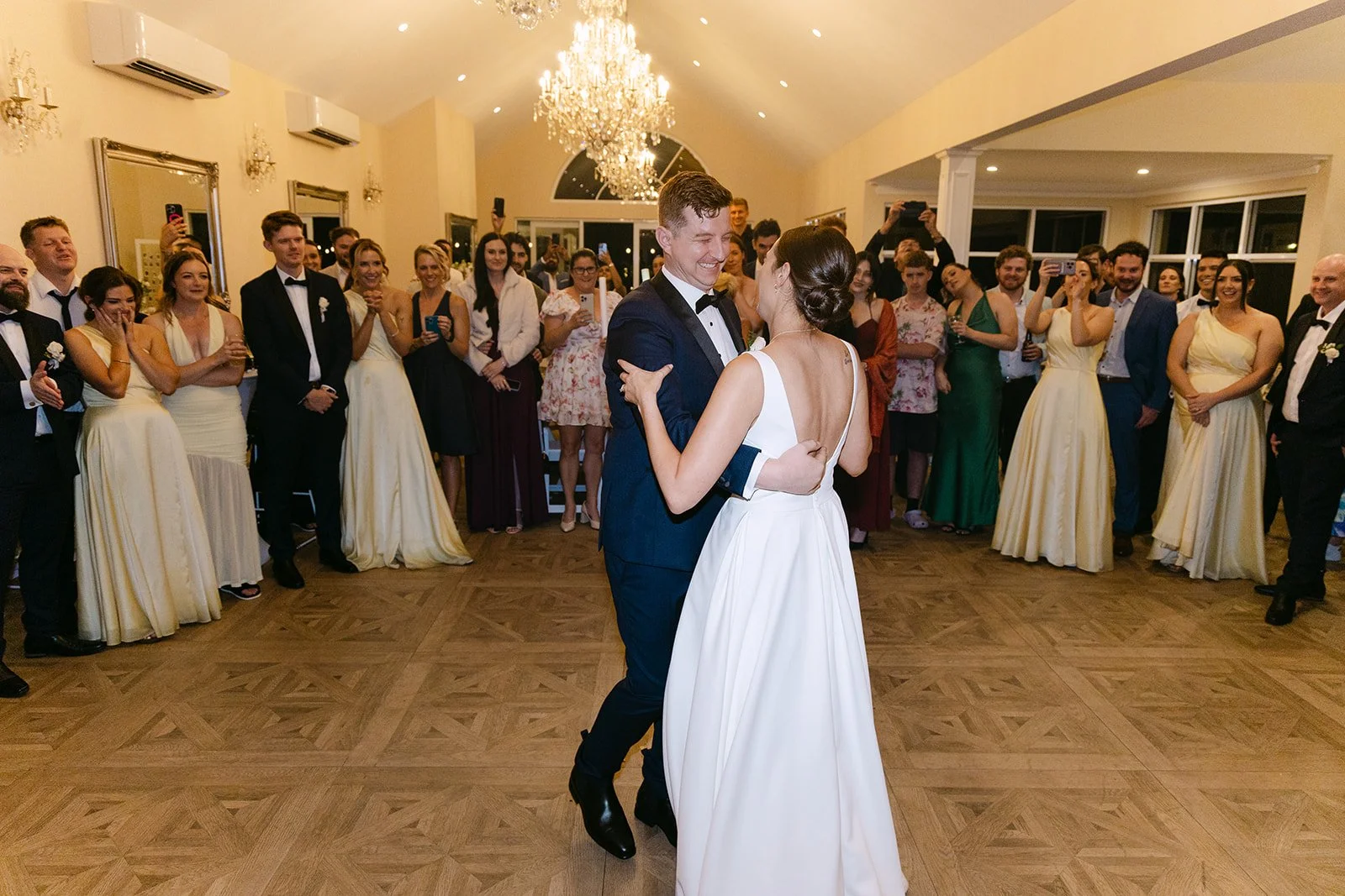A bride and groom dancing at their wedding reception with guests watching and taking photos in a beautifully decorated elegant ballroom.