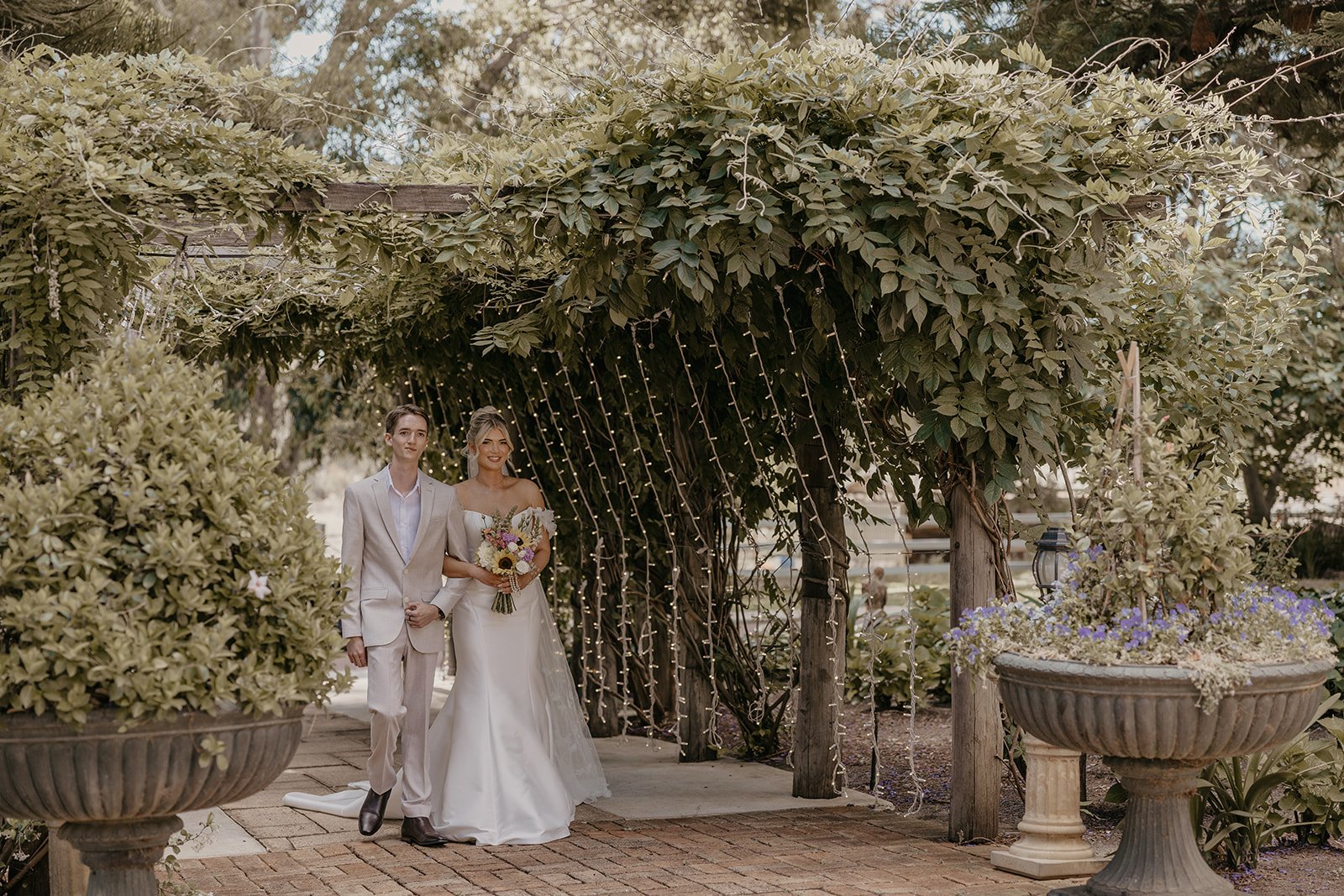 A bride and groom walking arm in arm under a decorated archway at their wedding, surrounded by potted plants and greenery.