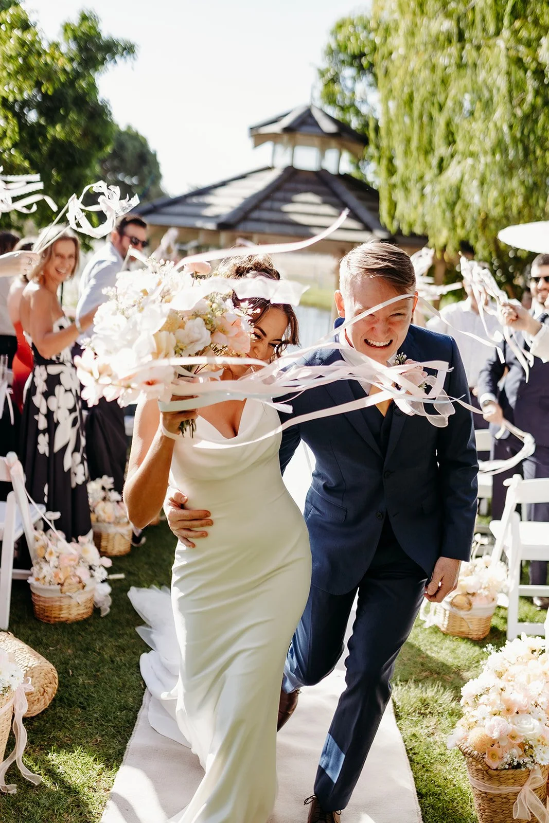 A bride and groom happily walk down the aisle at their outdoor wedding, surrounded by friends and family celebrating with confetti and flowers.