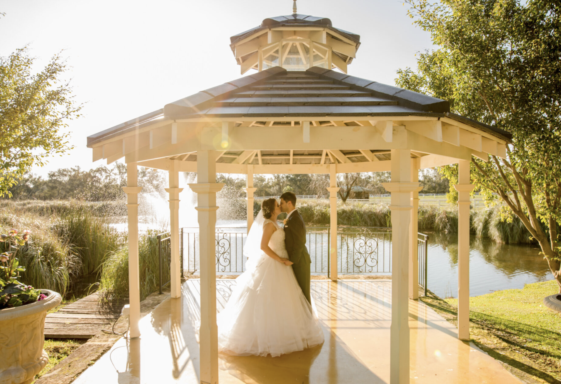 A bride and groom sharing a kiss under a gazebo in a garden during sunset.