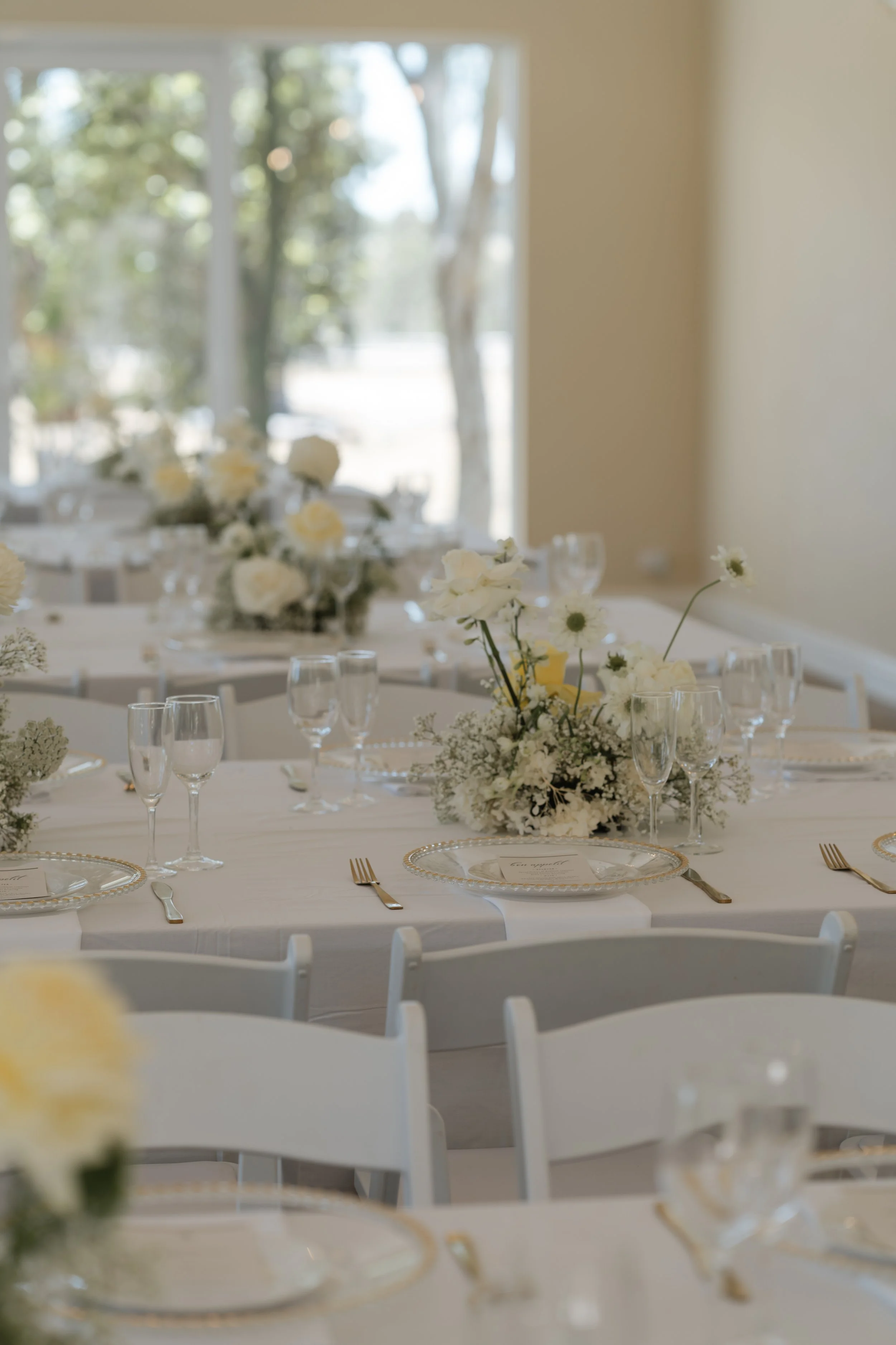 Elegant table setting with white floral centerpiece, glassware, and gold-rimmed plates in a bright room with large windows.