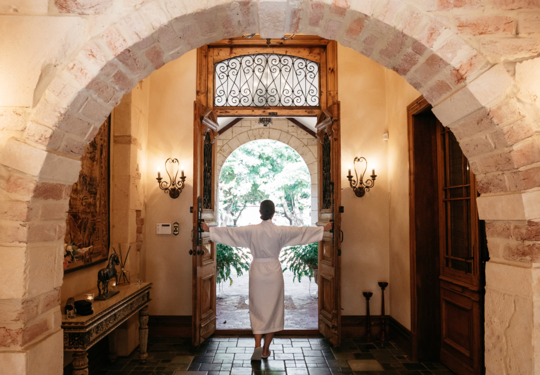 Person in a white bathrobe standing at an open wooden door, holding the door open and looking outside at lush greenery.