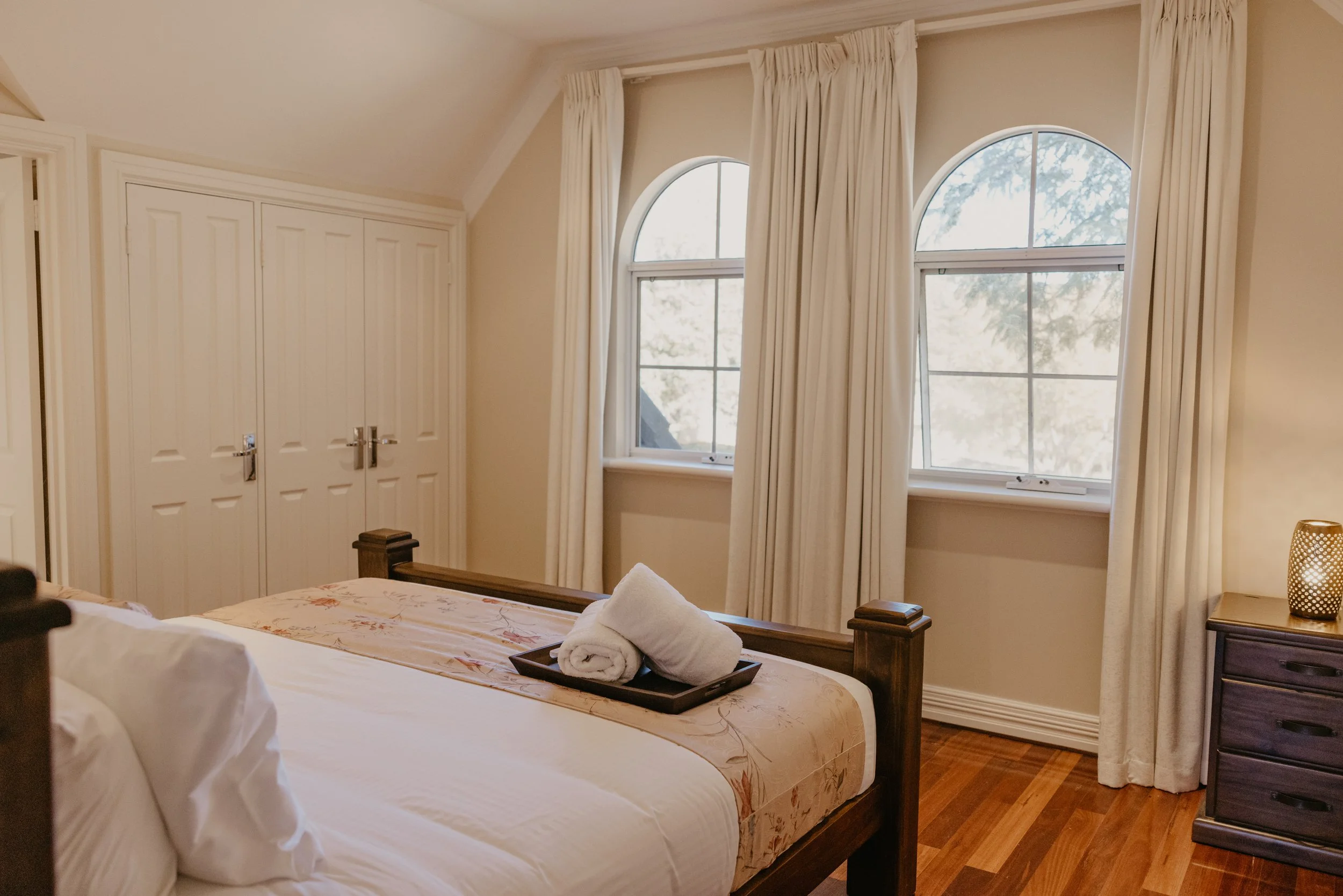 A bedroom with a wooden bed frame, white bedding, and freshly folded towels on the bed. There are two windows with arched tops and cream-colored curtains, and a dark wooden nightstand with a decorative lamp. The room has wood flooring and a built-in 